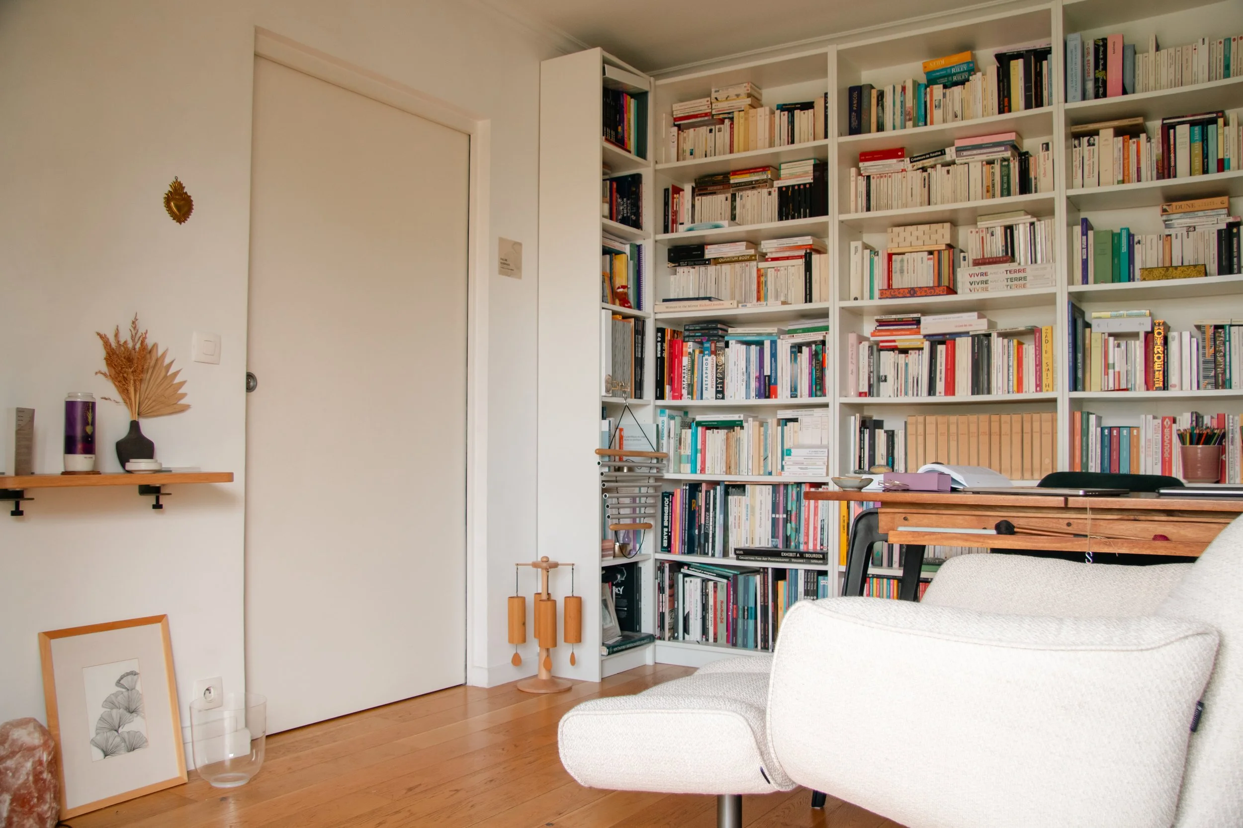 Une bibliothèque blanche remplie de livres avec une table en bois, une chaise en bois, et des décorations végétales sur un mur blanc. Le sol est en parquet en bois clair.