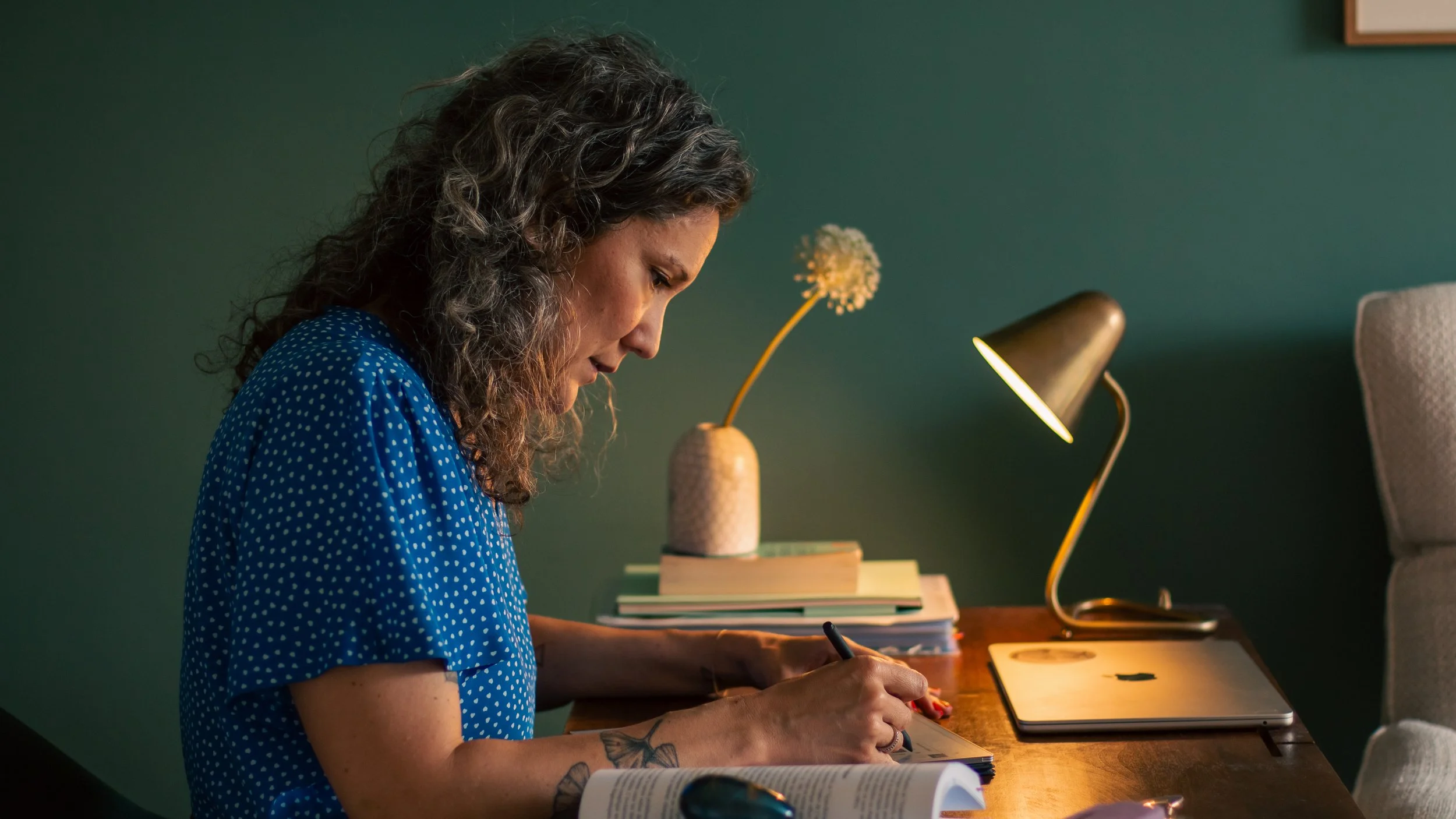 Une femme aux cheveux bouclés et portant une robe bleue à pois travaille à un bureau, avec une lampe de bureau, des livres et un ordinateur portable.