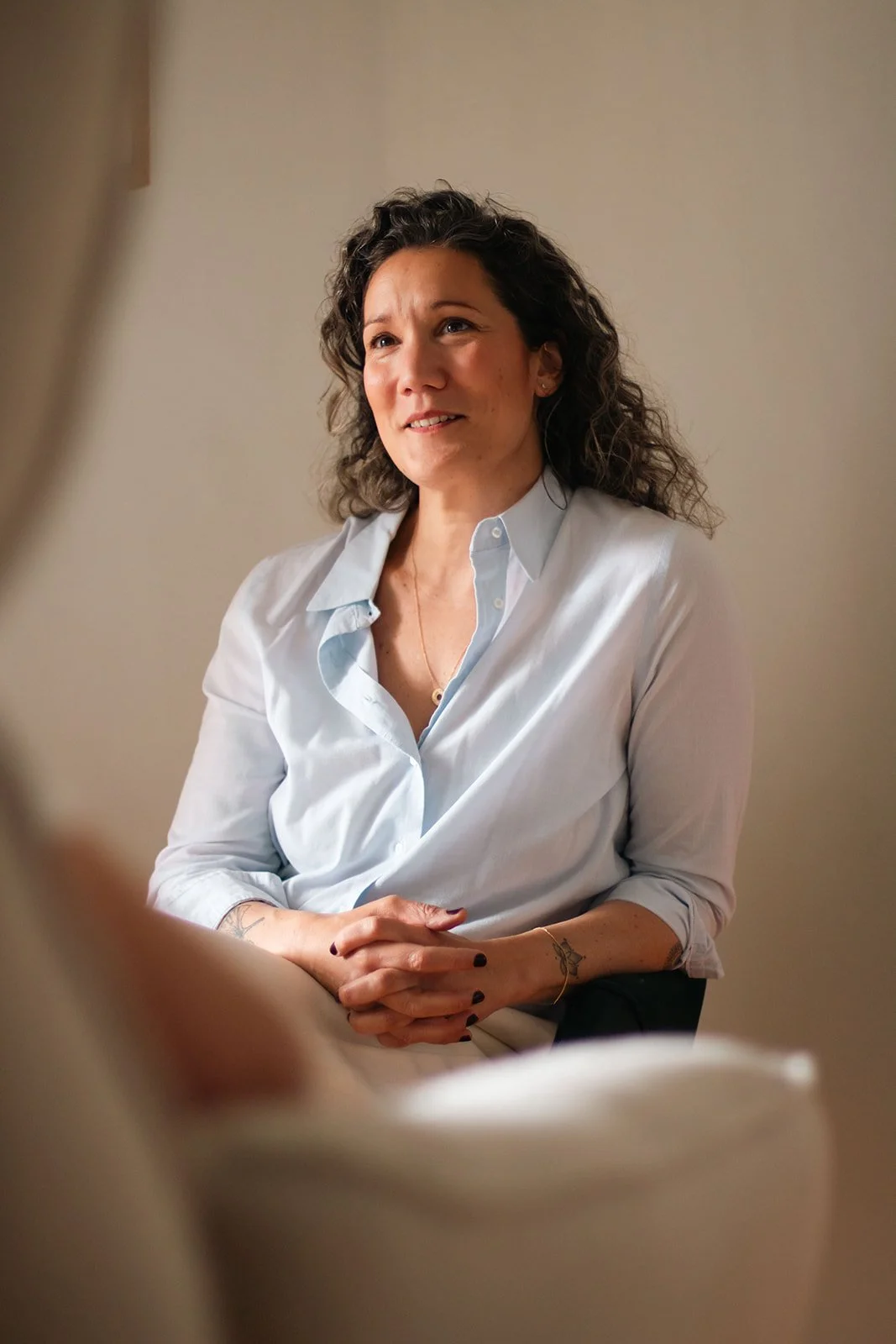 Une femme aux cheveux bouclés, portant une chemise blanche, semble converser ou écouter attentivement, avec un sourire léger, dans un environnement intérieur.