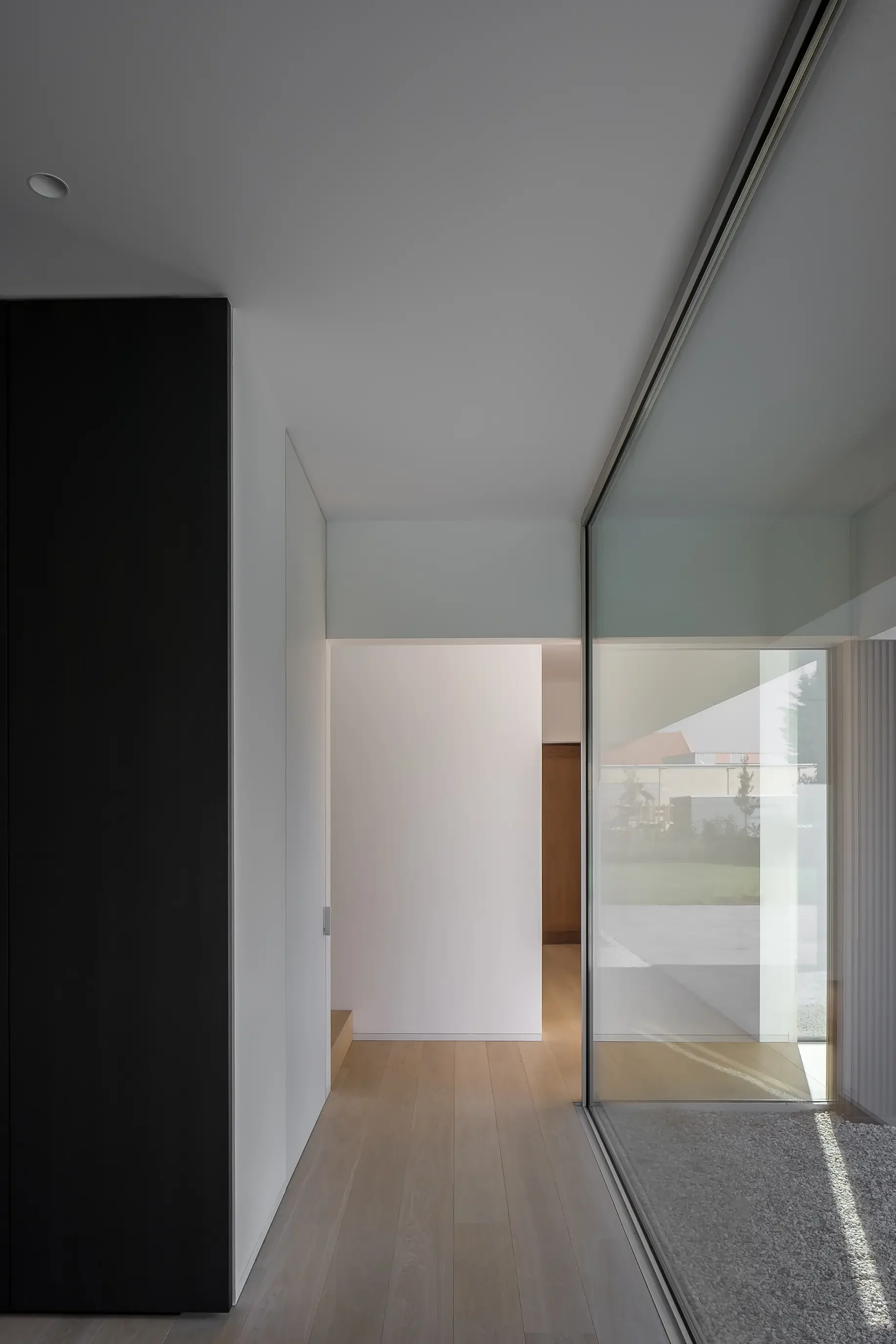 Interior of a modern home with light wooden flooring, a black wall on the left, and a large glass sliding door on the right leading outside. The ceiling is white with an inset light and a track for a door or window system.