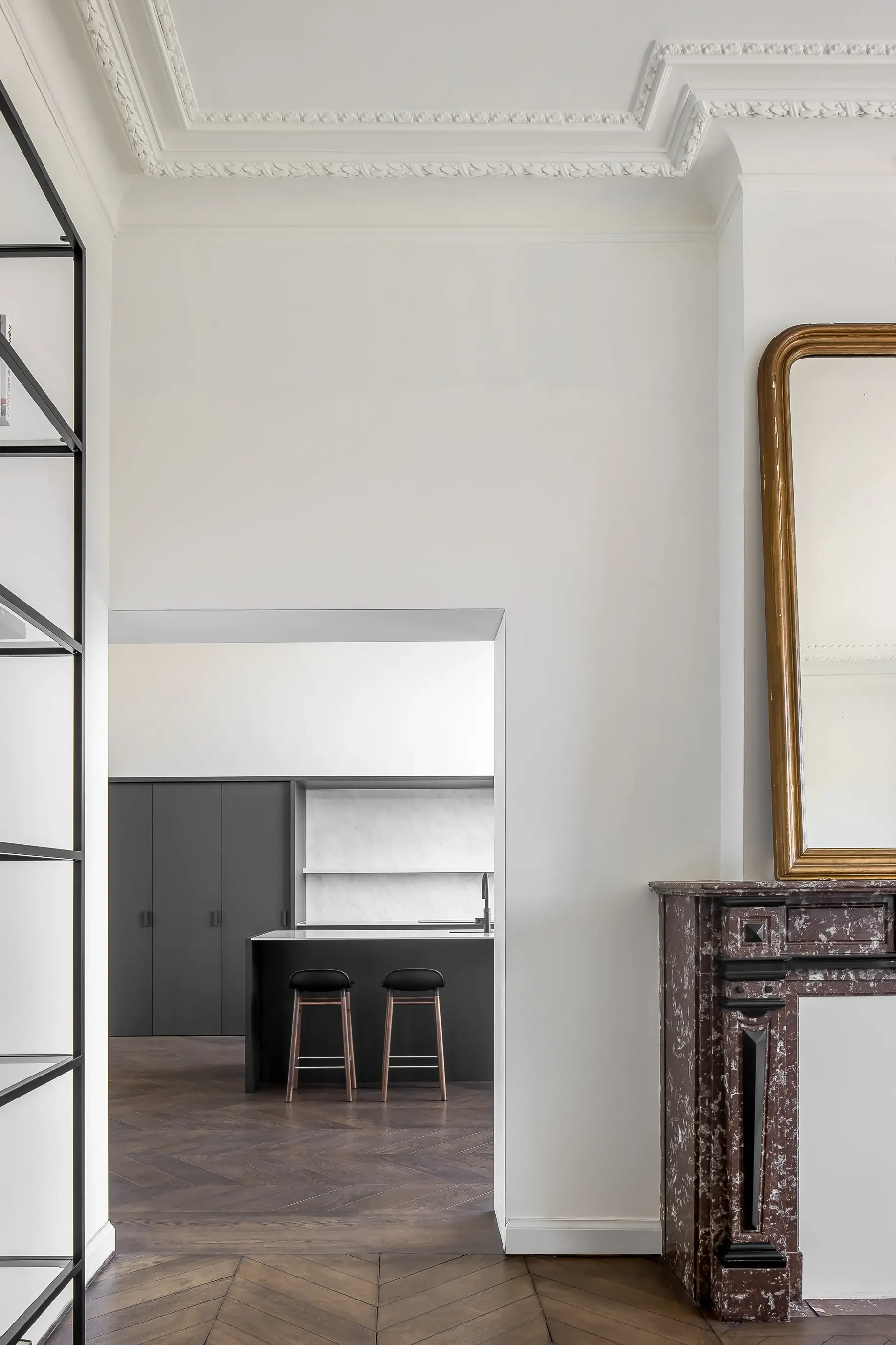 Interior room with ornate white ceiling molding, a partial view of a mirror frame, a marble fireplace, a small kitchen or bar area with two stools, and wood flooring.