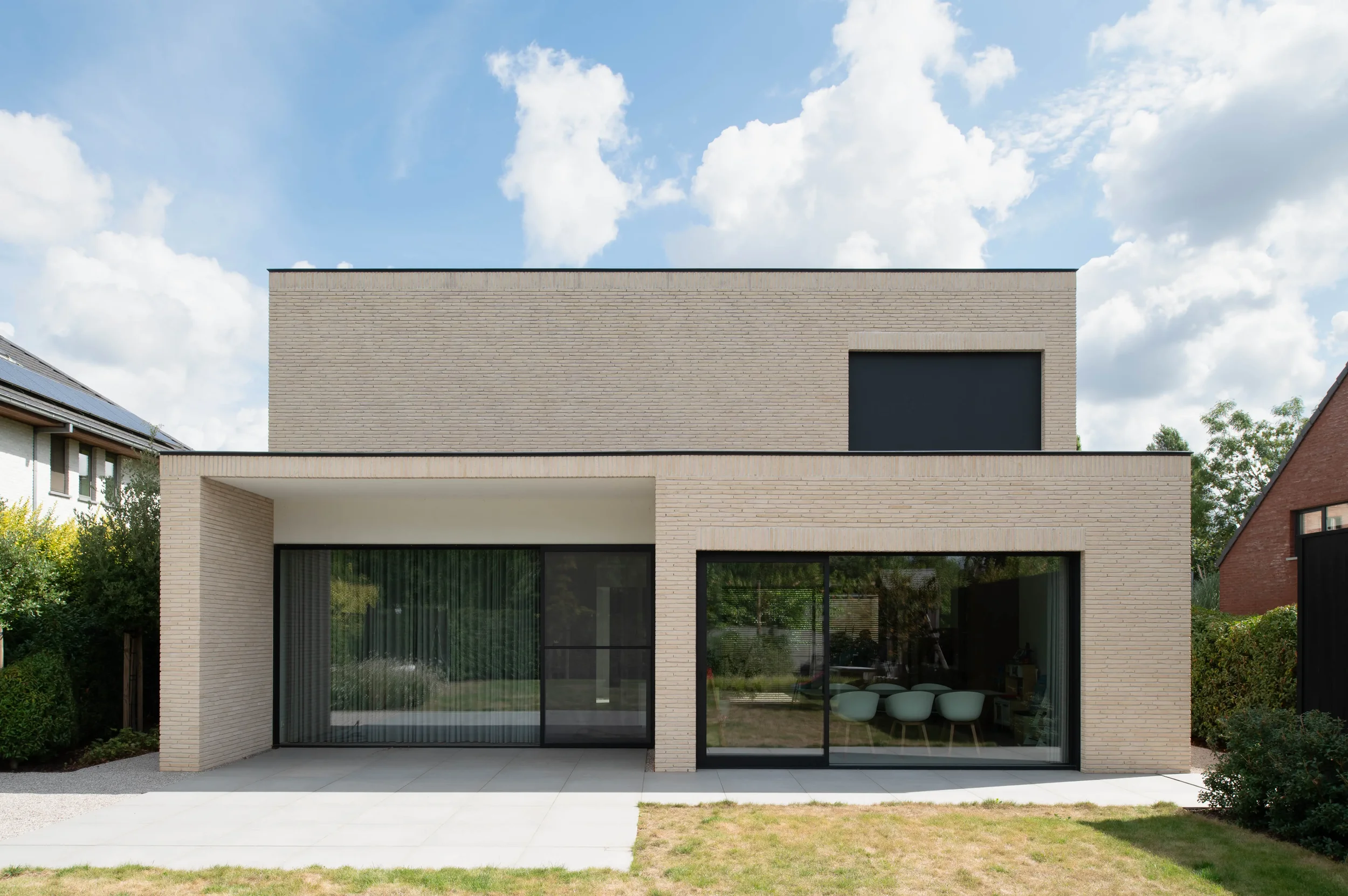 Modern two-story house with large glass sliding doors, light-colored brick exterior, and patio area, under a partly cloudy sky.