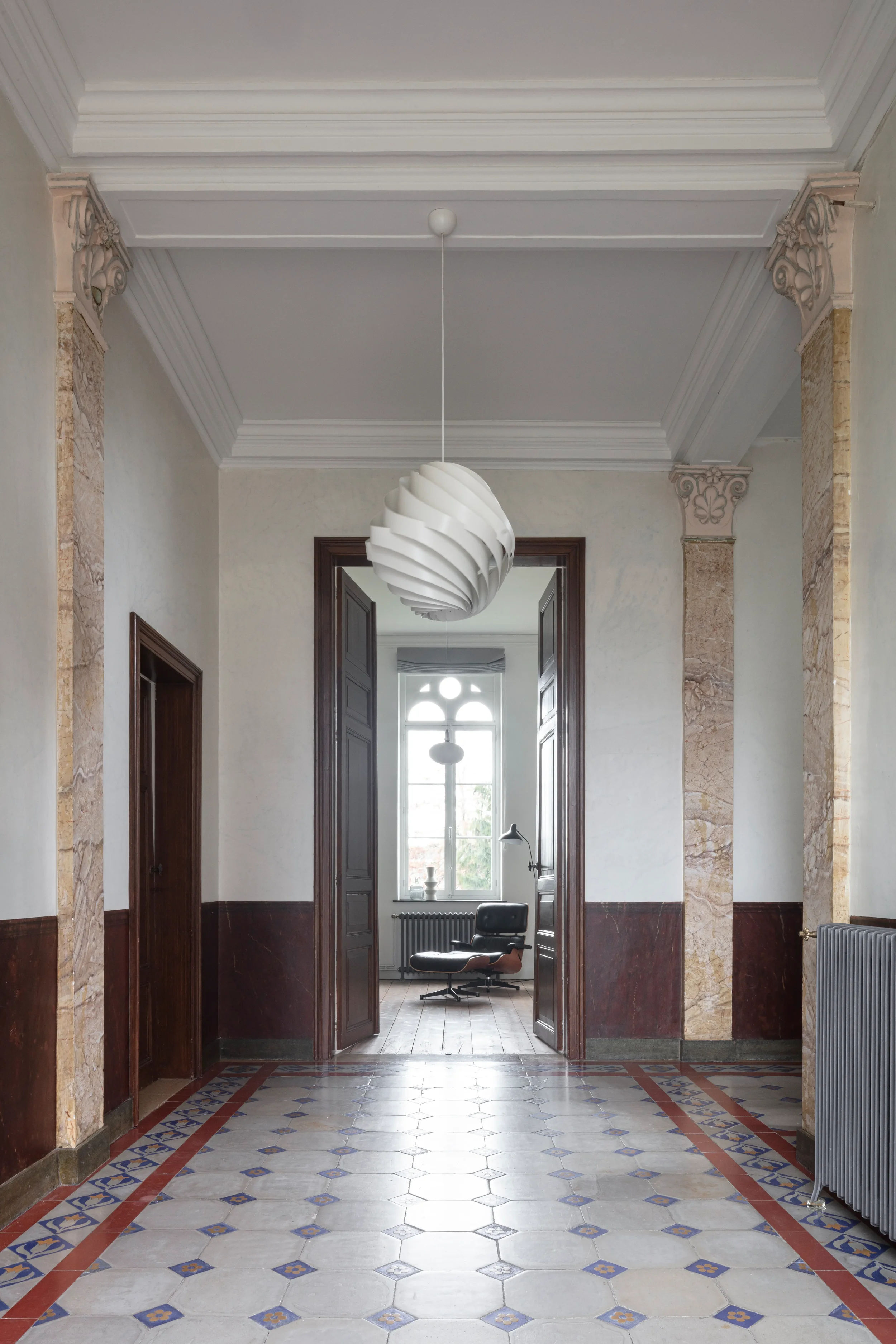Interior view of a hallway with a patterned tiled floor, ornate columns and trim, a modern pendant light fixture hanging from the ceiling, and a room with a window and furniture visible through double doors.