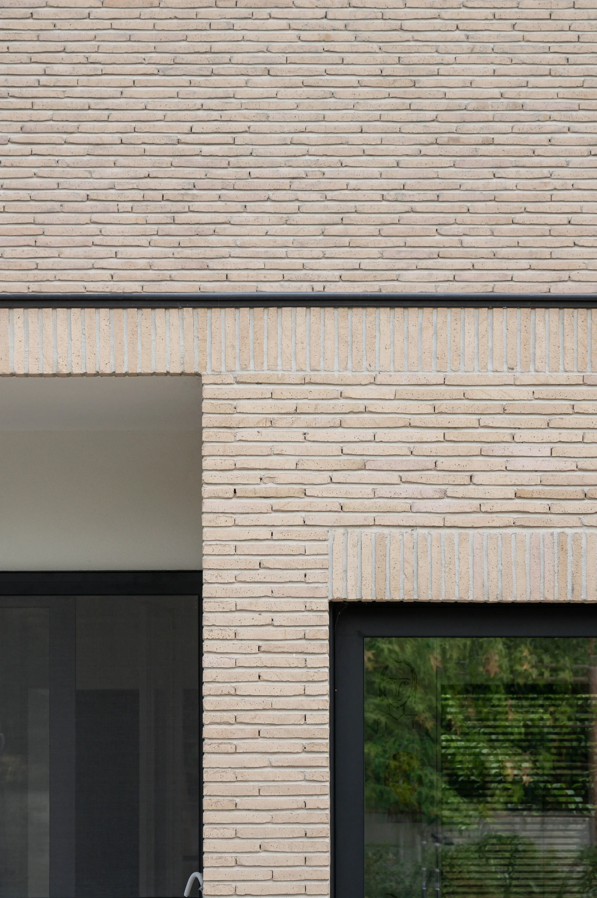 Exterior view of a modern building with beige brick walls, black window frames, and greenery visible through the window.