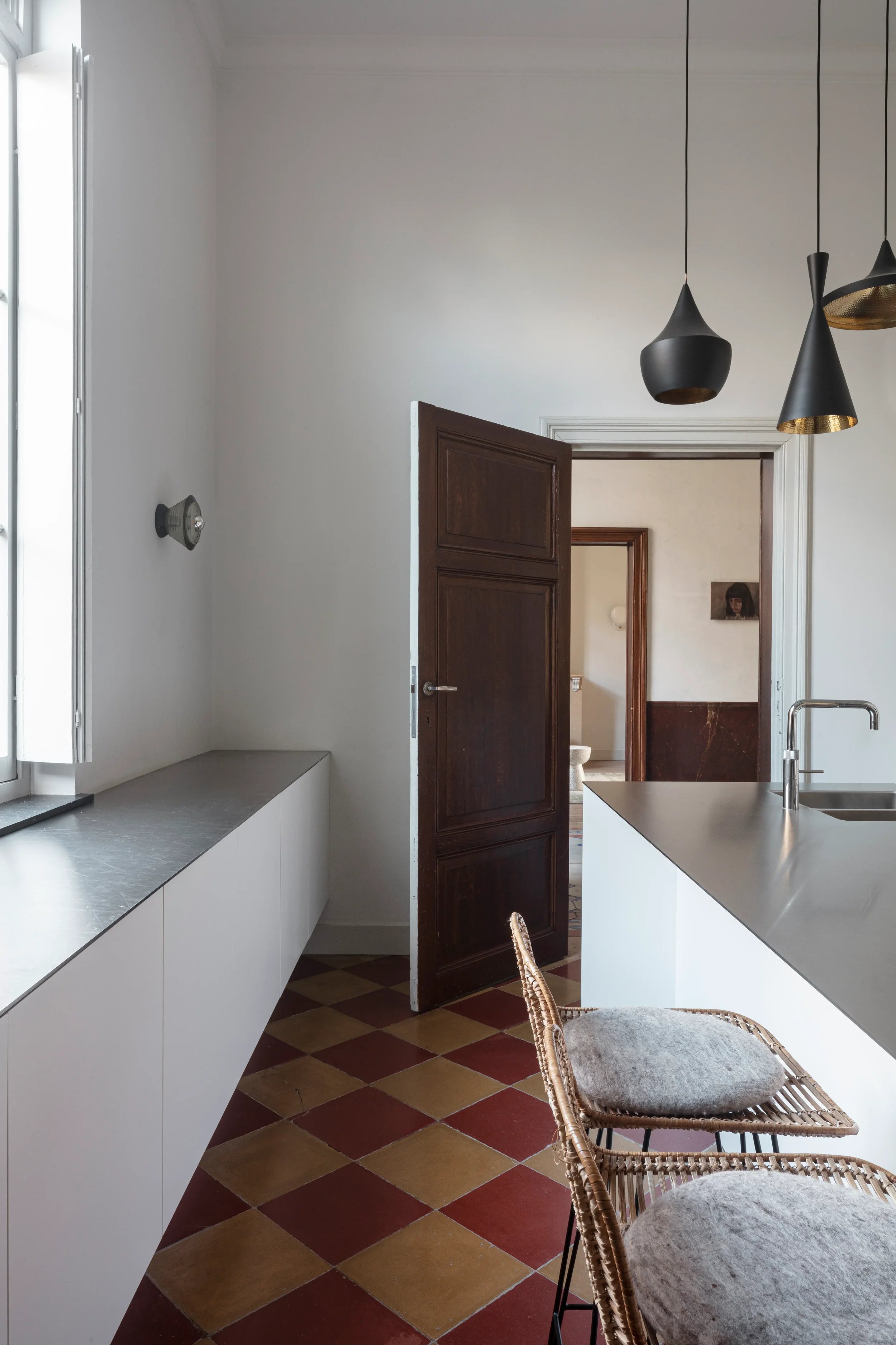 A modern kitchen with white cabinets and a gray countertop. A wicker chair with a cushioned seat is in the foreground. The brick floor has a red and yellow checkerboard pattern. A doorway leads to a room with a small picture of a woman with dark hair on the wall.