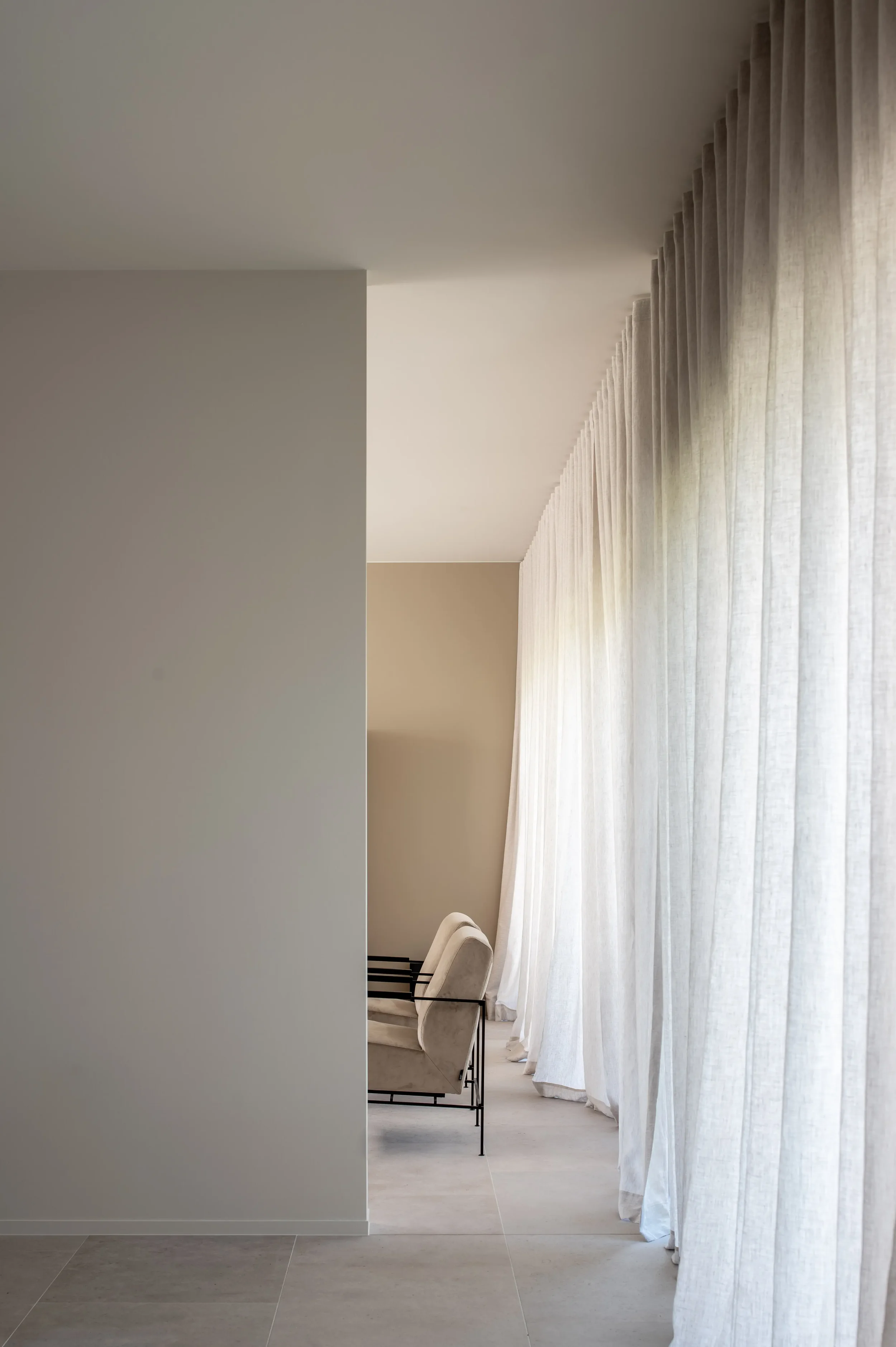 A corner of a room with a light-colored wall and a large window with sheer white curtains, a plush armchair, and a light-colored tiled floor.
