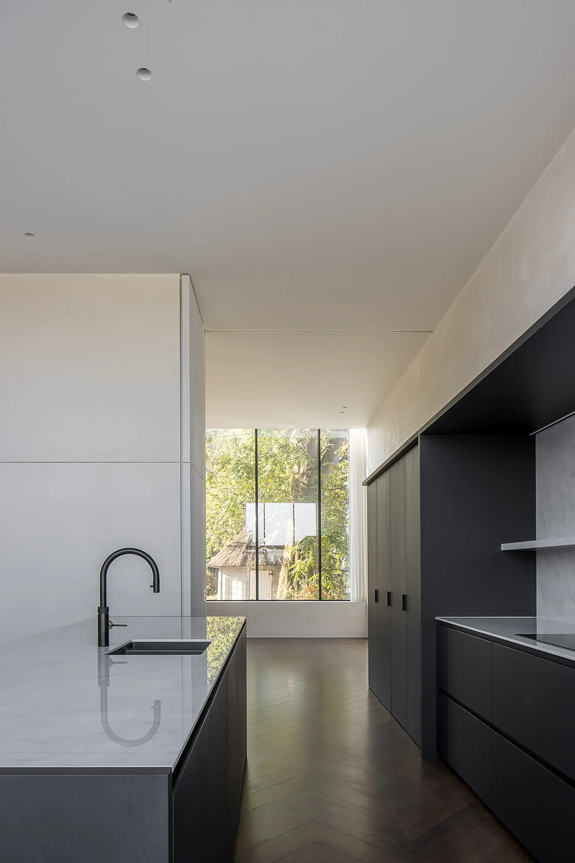 Modern kitchen with white countertops, a black sink and faucet, dark gray cabinets, large window showing trees, and a light-colored wooden floor.