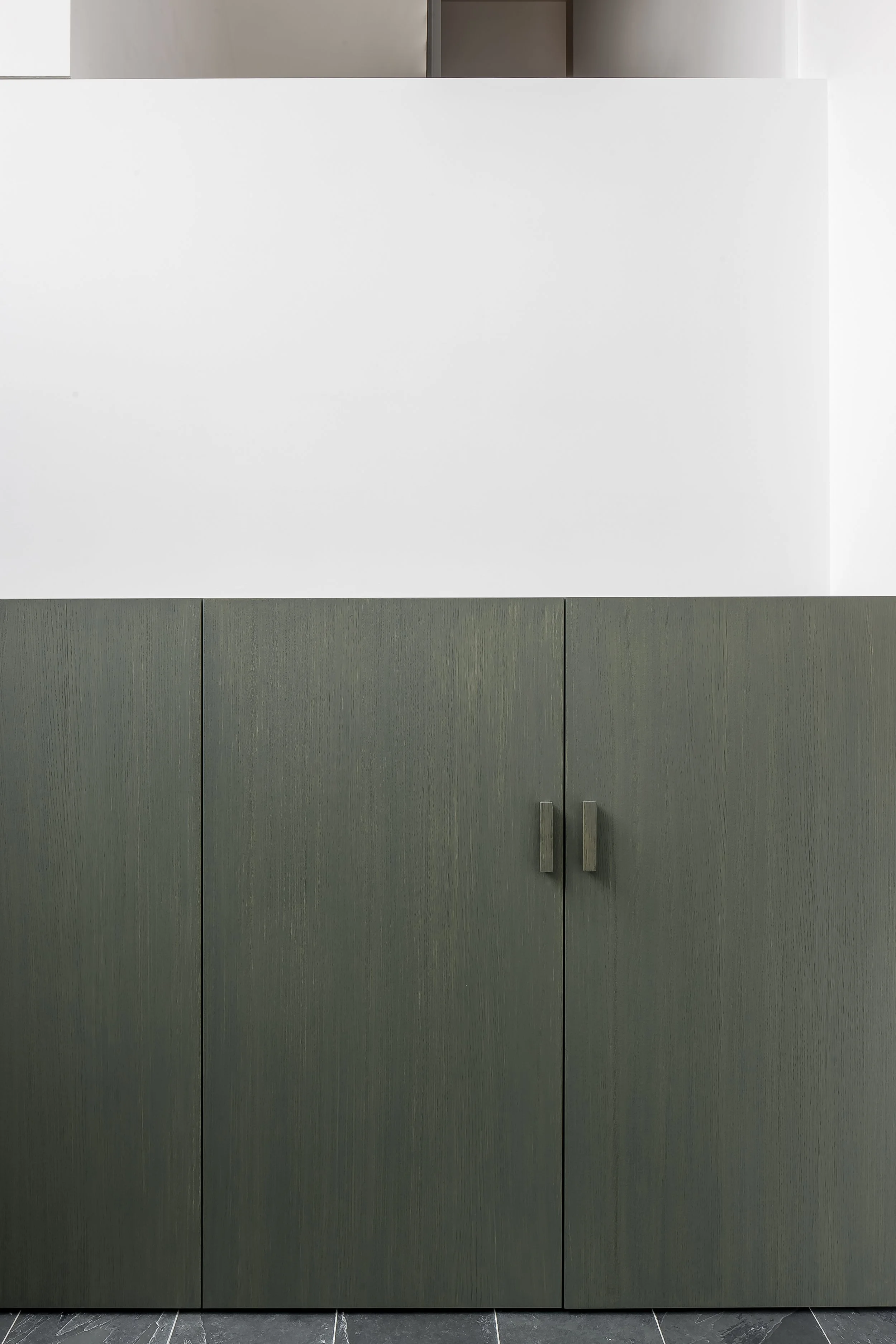 A gray double-door cabinet with two handles, positioned below a white wall and a gray upper cabinet, on a dark tiled floor.