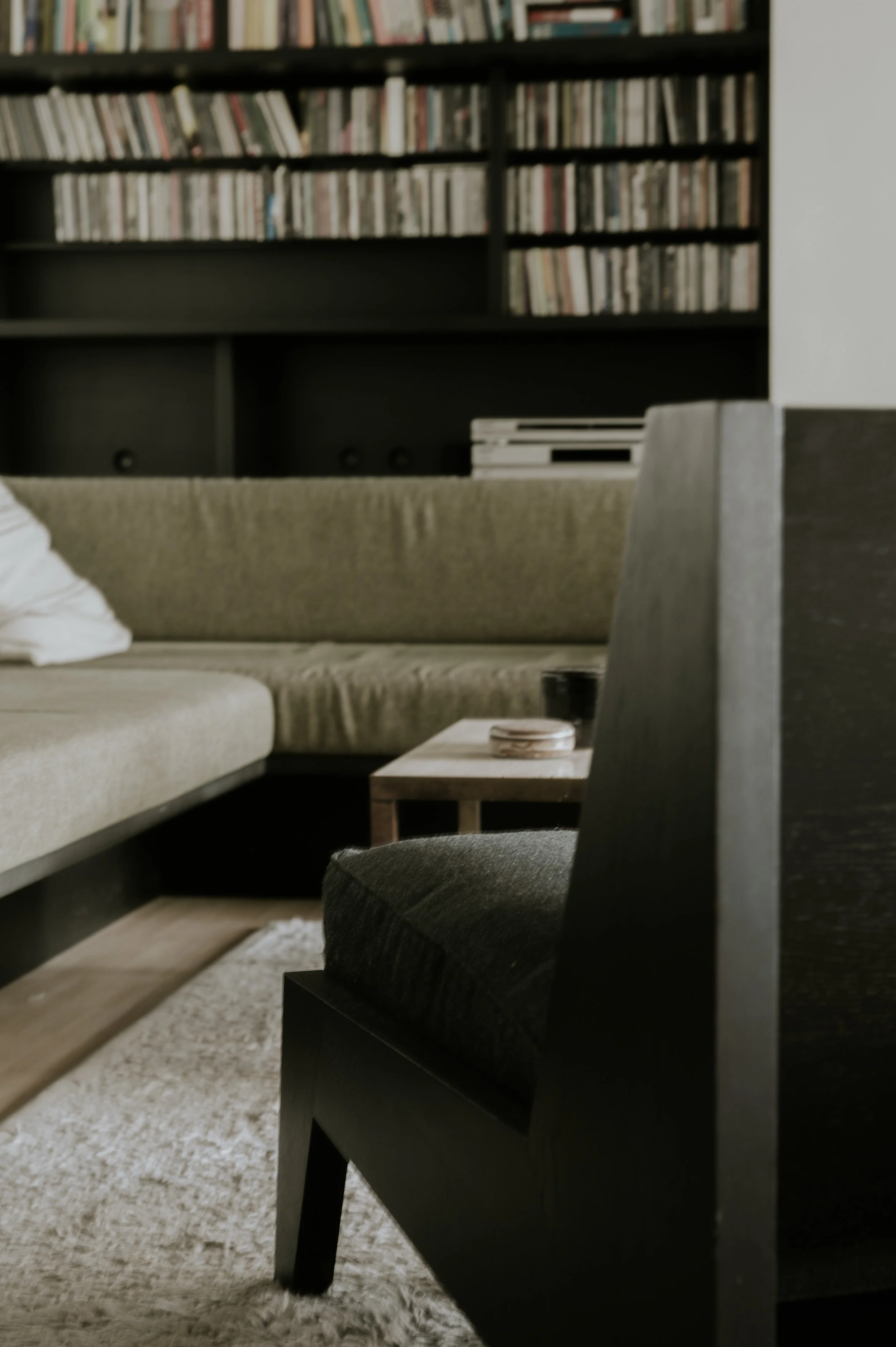 A living room with a black armchair in the foreground, a beige couch with a pillow, a wooden side table with a coaster, a carpet, and a bookshelf filled with books in the background.