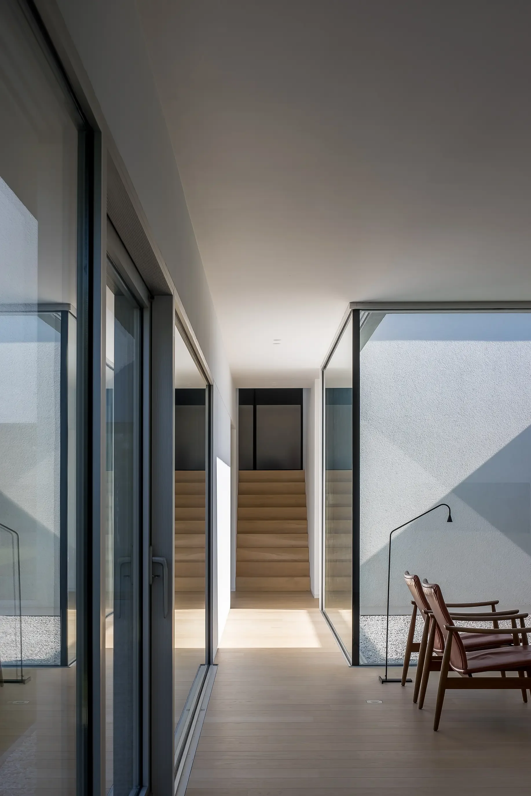 Modern indoor space with large glass sliding doors, wooden stairs, white walls, and outdoor patio seating with chairs and a wall-mounted lamp.