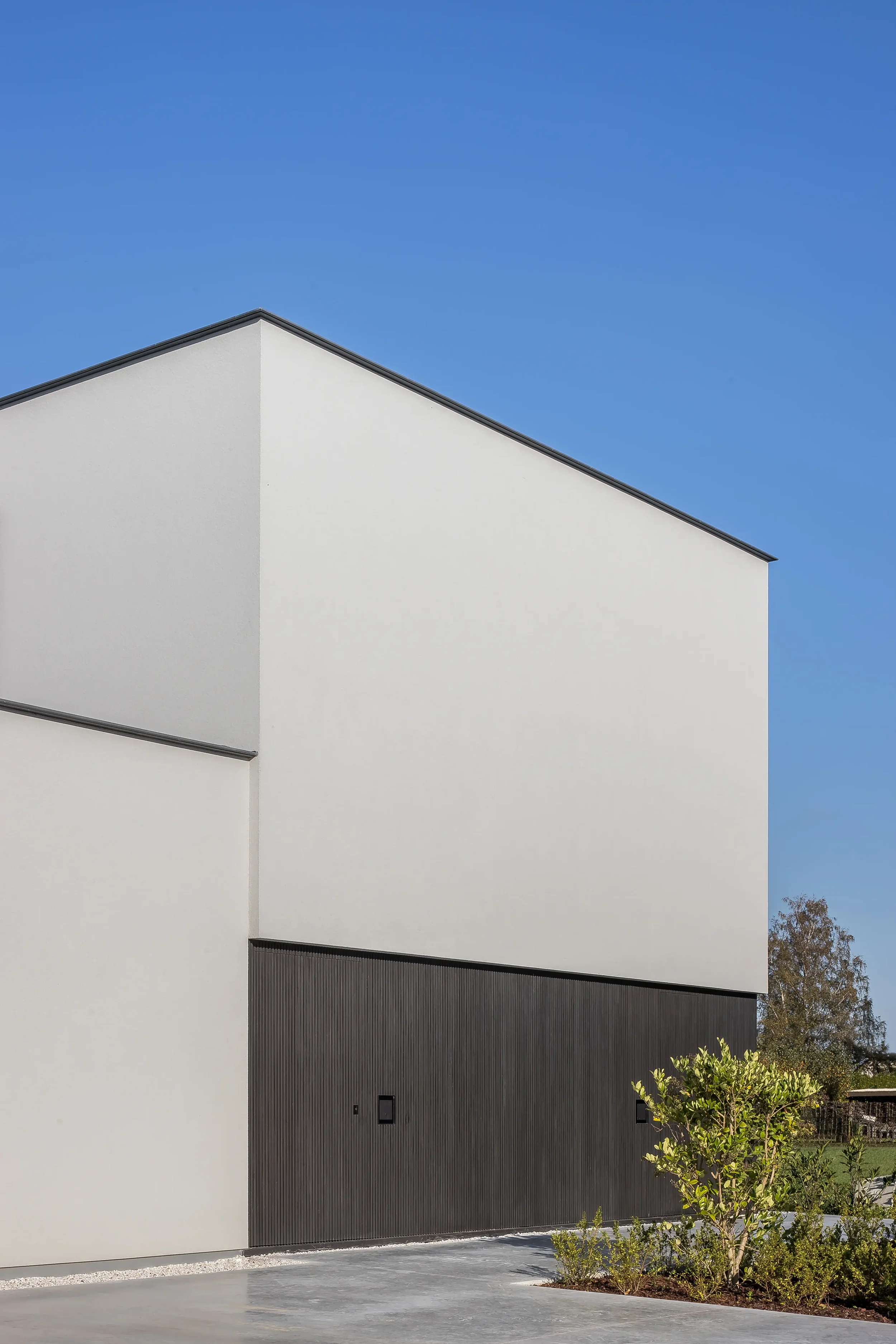 Modern building with white walls and black garage door, small trees, and clear blue sky.