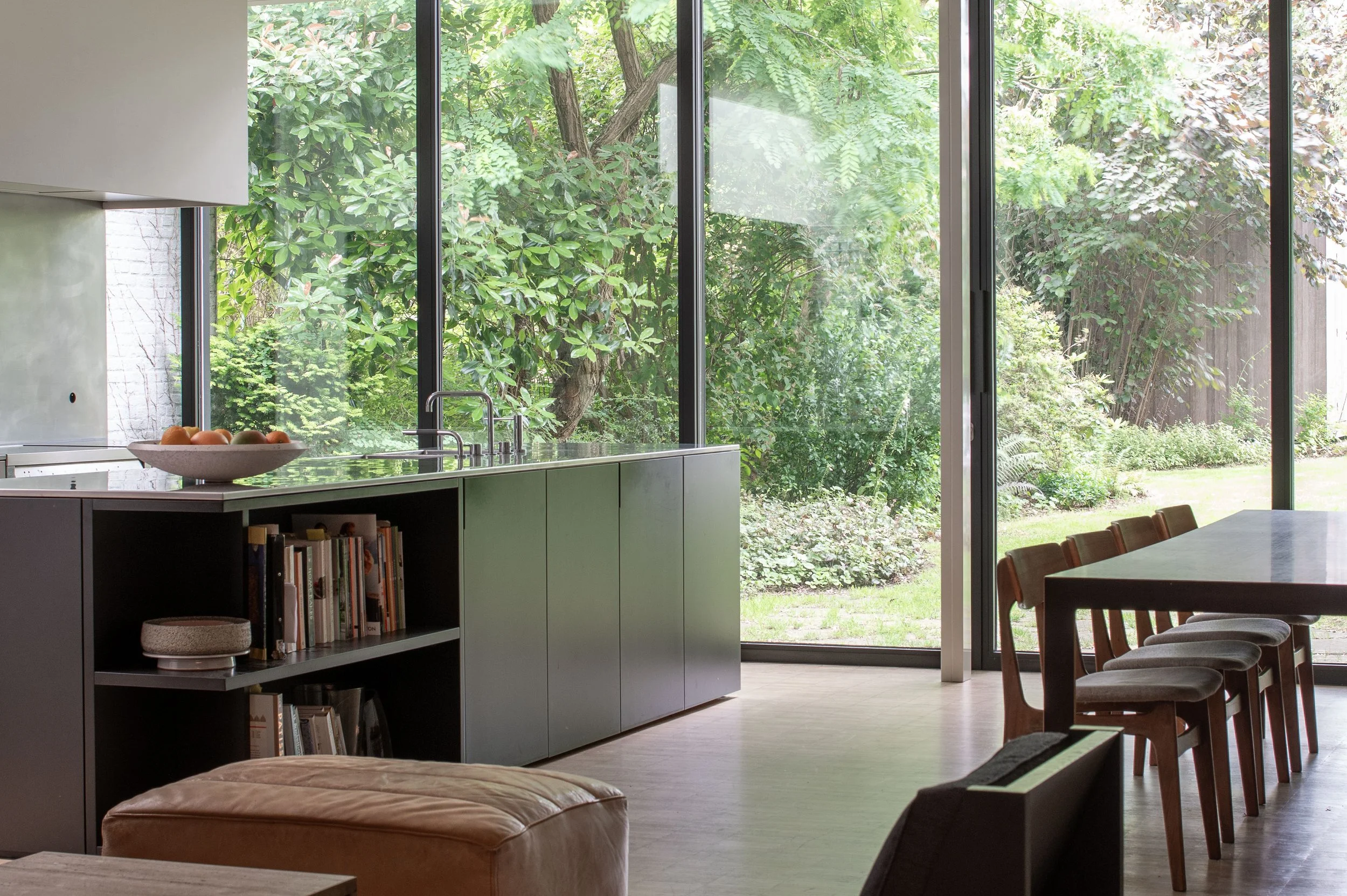 Modern dining room with a large glass window overlooking a lush green garden with trees and bushes. The room features a long wooden dining table with chairs, a bookshelf with books, and a beige ottoman.