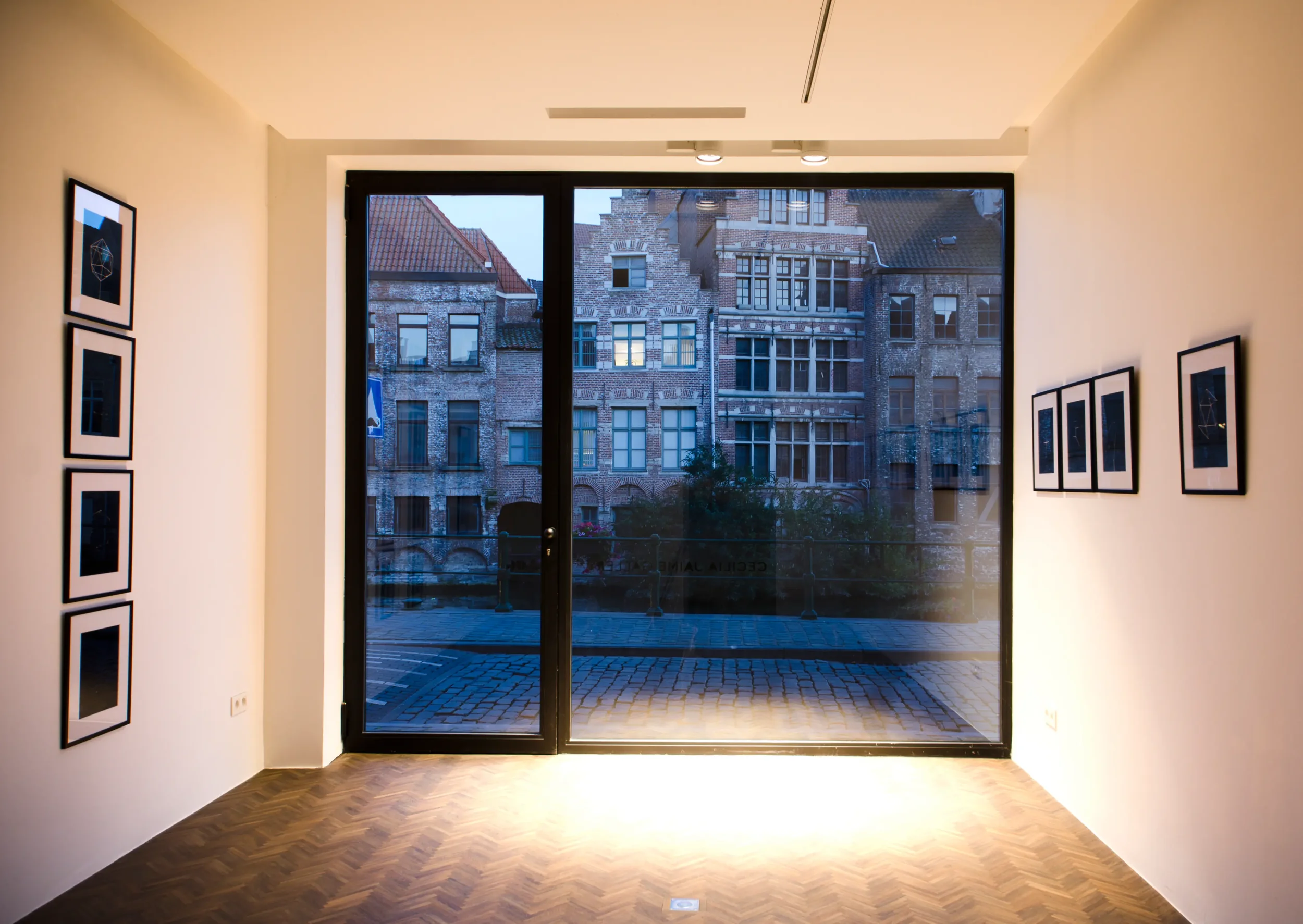 Interior view of a gallery or exhibition space with framed artwork on white walls, large glass door leading outside, and a view of historic brick buildings with tiled roofs across the street.