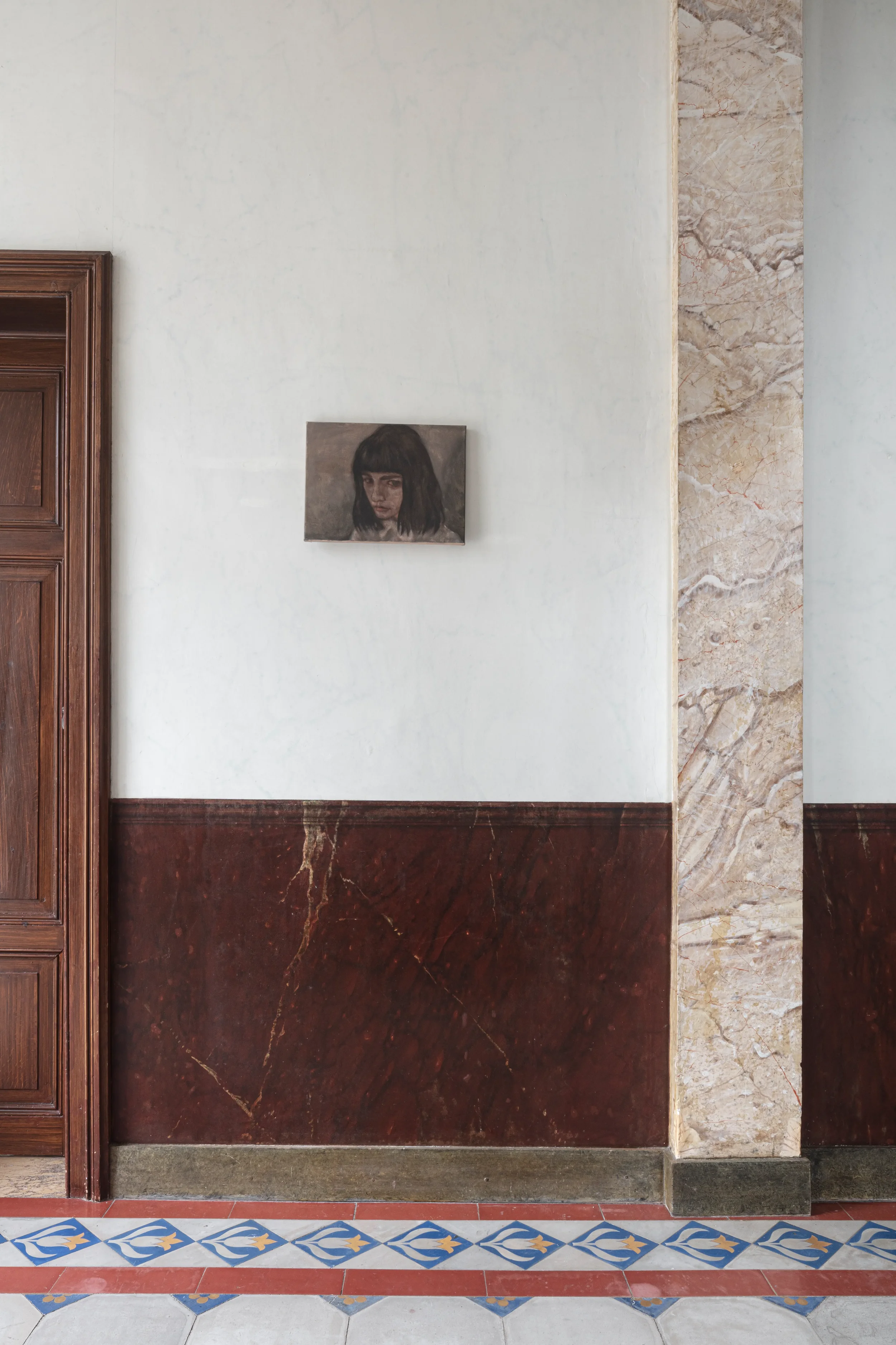 Interior wall decor with small portrait painting of a woman with dark hair, decorative mosaic tiles on the floor, and a wooden doorframe.
