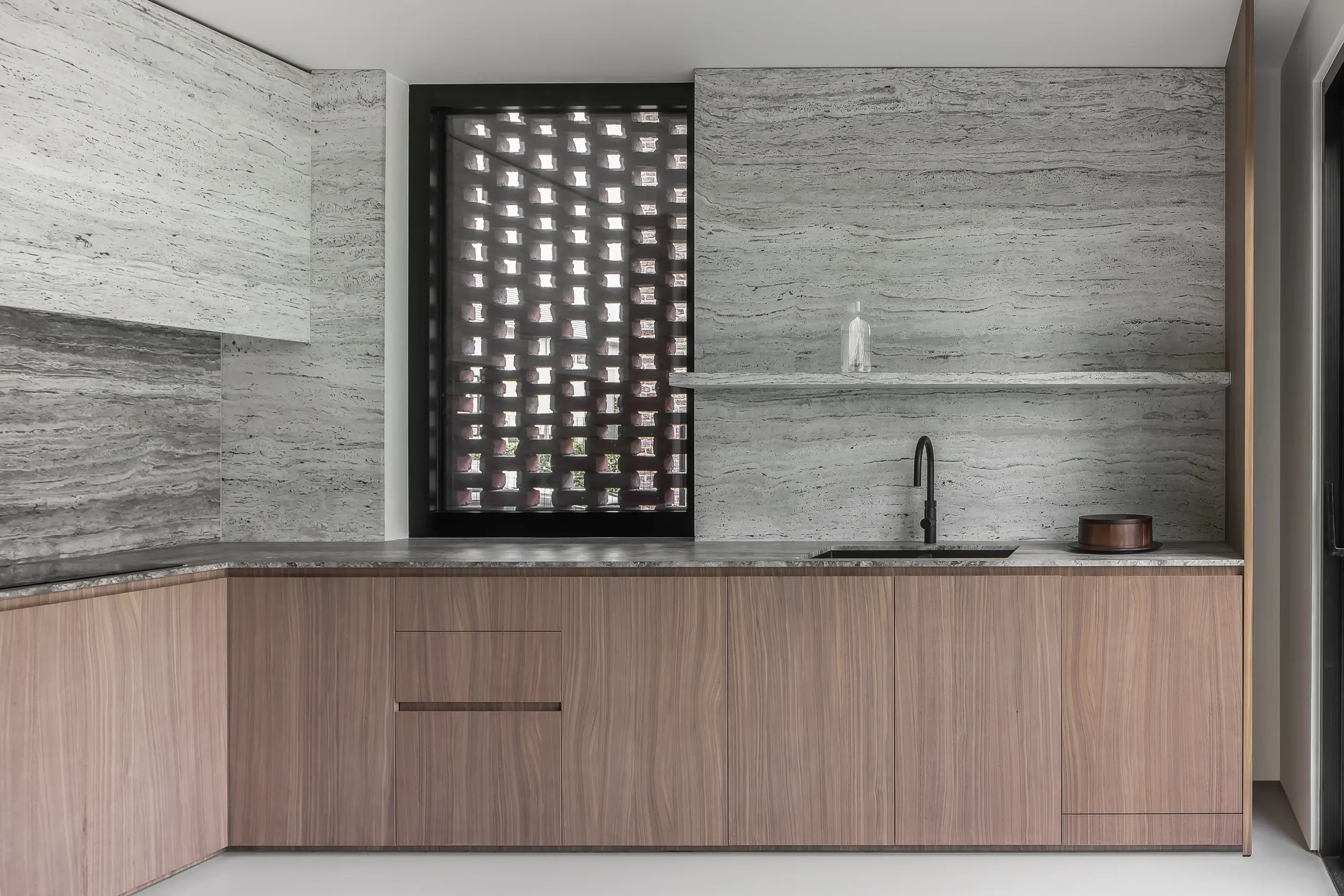 Modern minimalist kitchen with light gray marble countertops and backsplash, wooden cabinets, black faucet, a glass bottle, and a decorative wooden bowl.