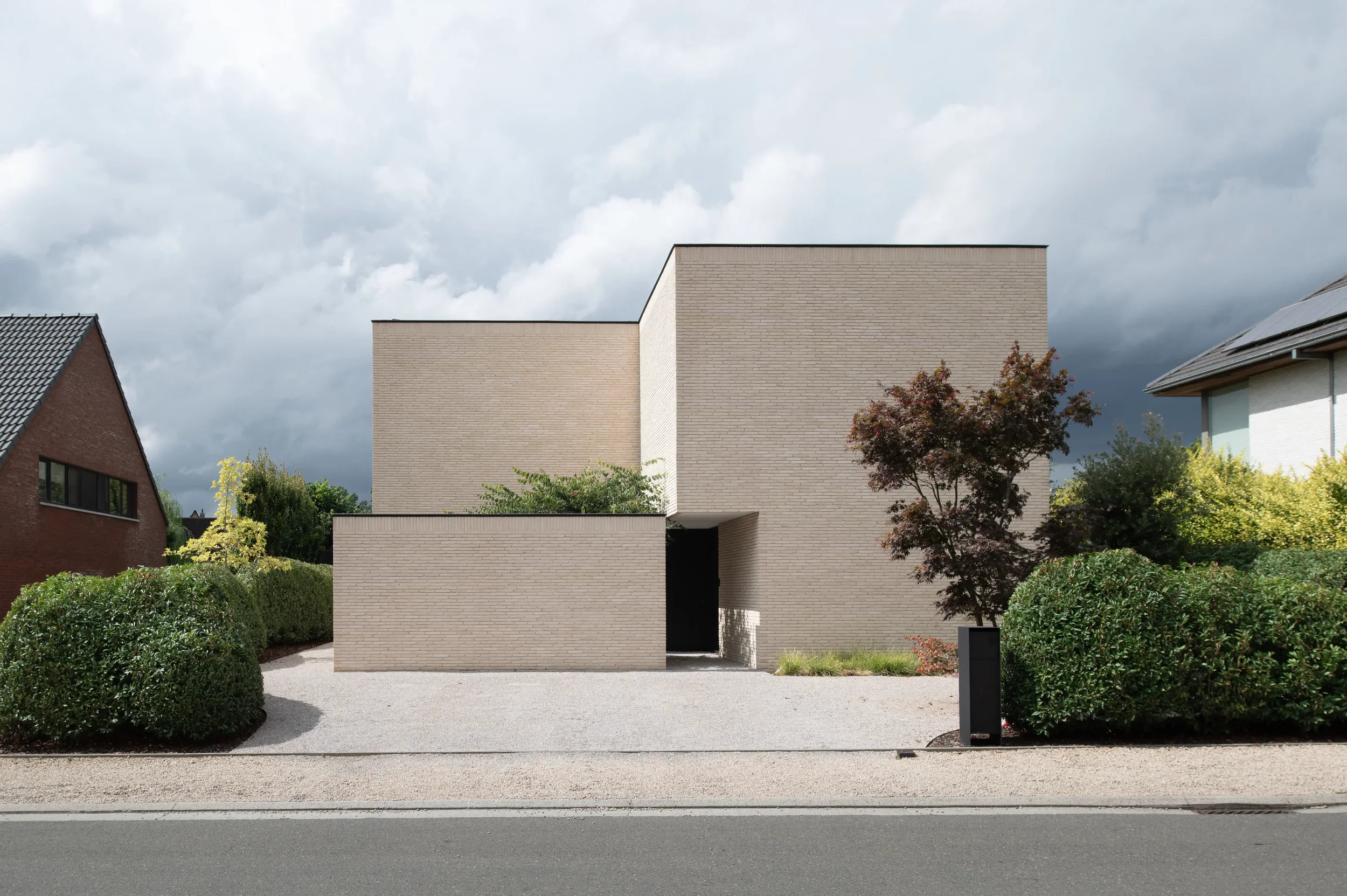Modern white brick house with clean lines, dark entrance, surrounded by greenery, under a cloudy sky.