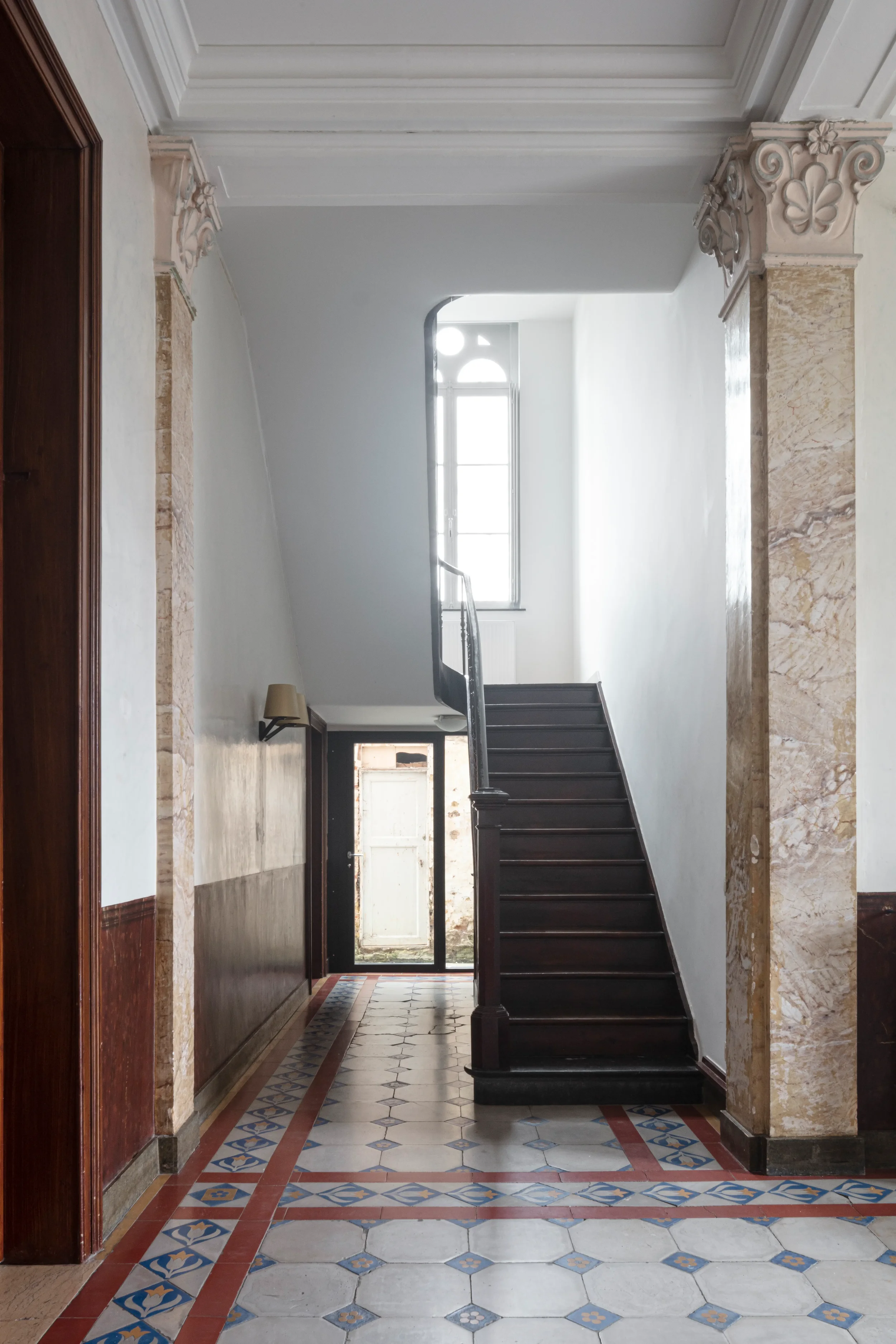 Interior of a hallway with a staircase, patterned tile floor, and a window at the top of the stairs allowing natural light
