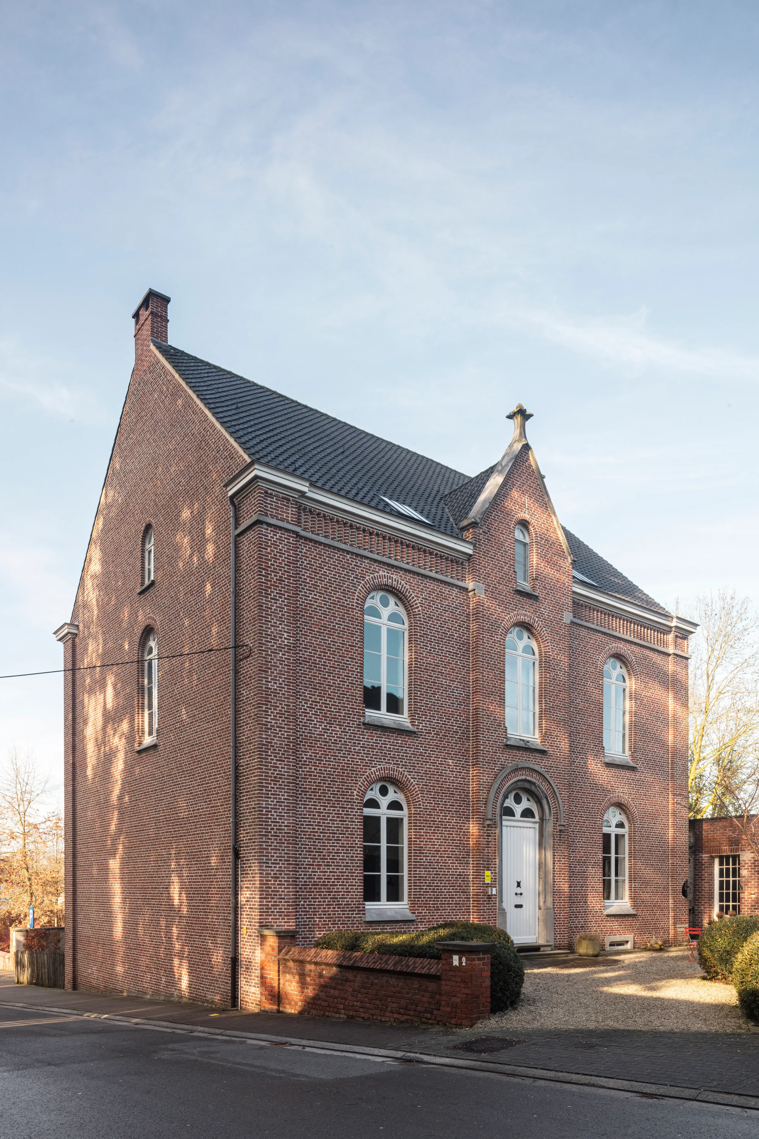 A red brick building with arched windows and a white door, surrounded by a brick wall and a small gravel yard, under a clear blue sky.