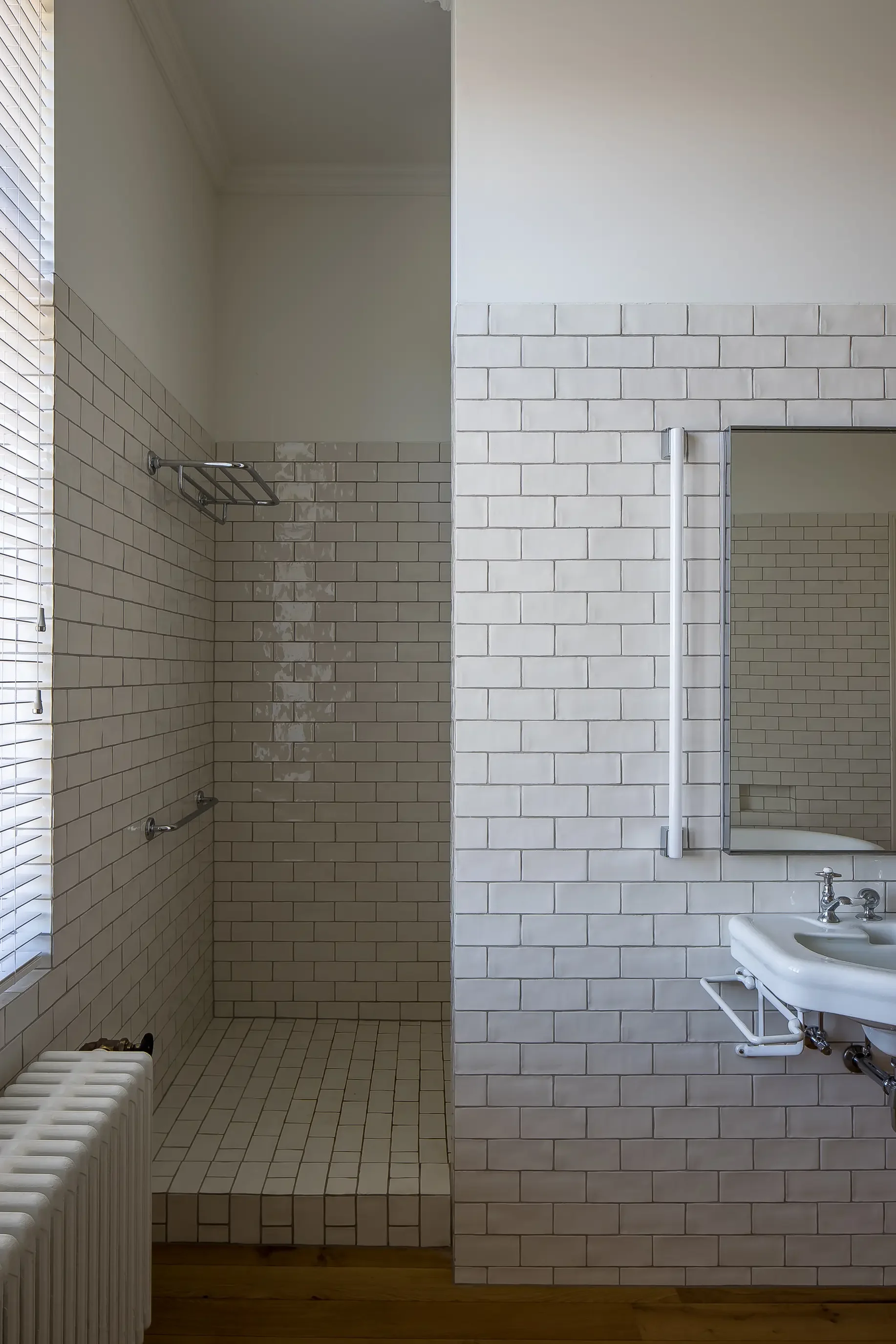 A bathroom with beige tiles on the floor and walls, a small white sink with a silver faucet, a mirror with a white frame, a towel bar, and a radiator. The left side shows a window with blinds and a shower area with a towel rack.