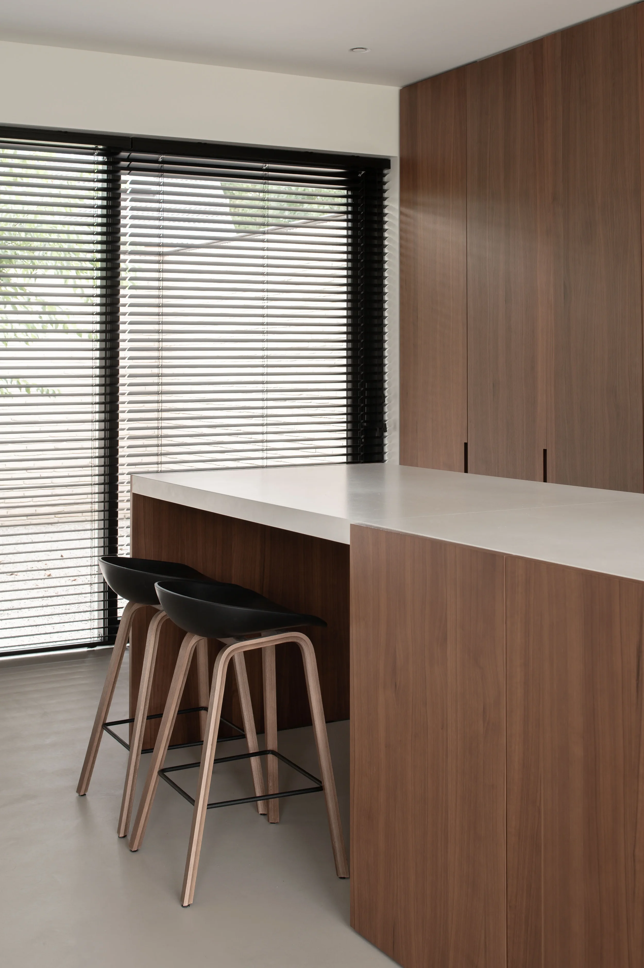 Modern kitchen with a white countertop, wooden cabinetry, black bar stools, and large windows with black blinds.