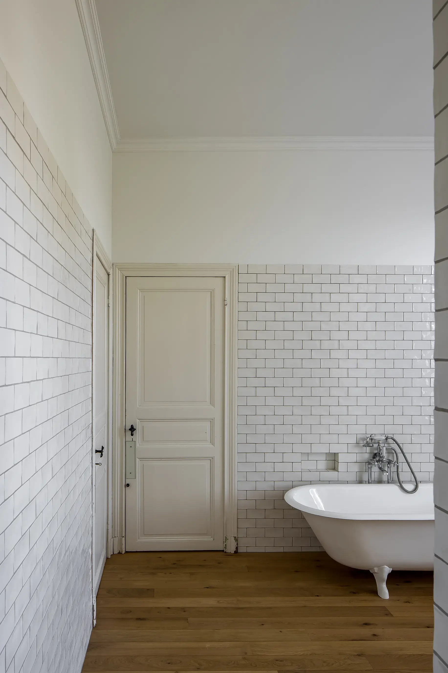 A bathroom with white subway tile walls, a vintage clawfoot bathtub, wood flooring, and a white door.