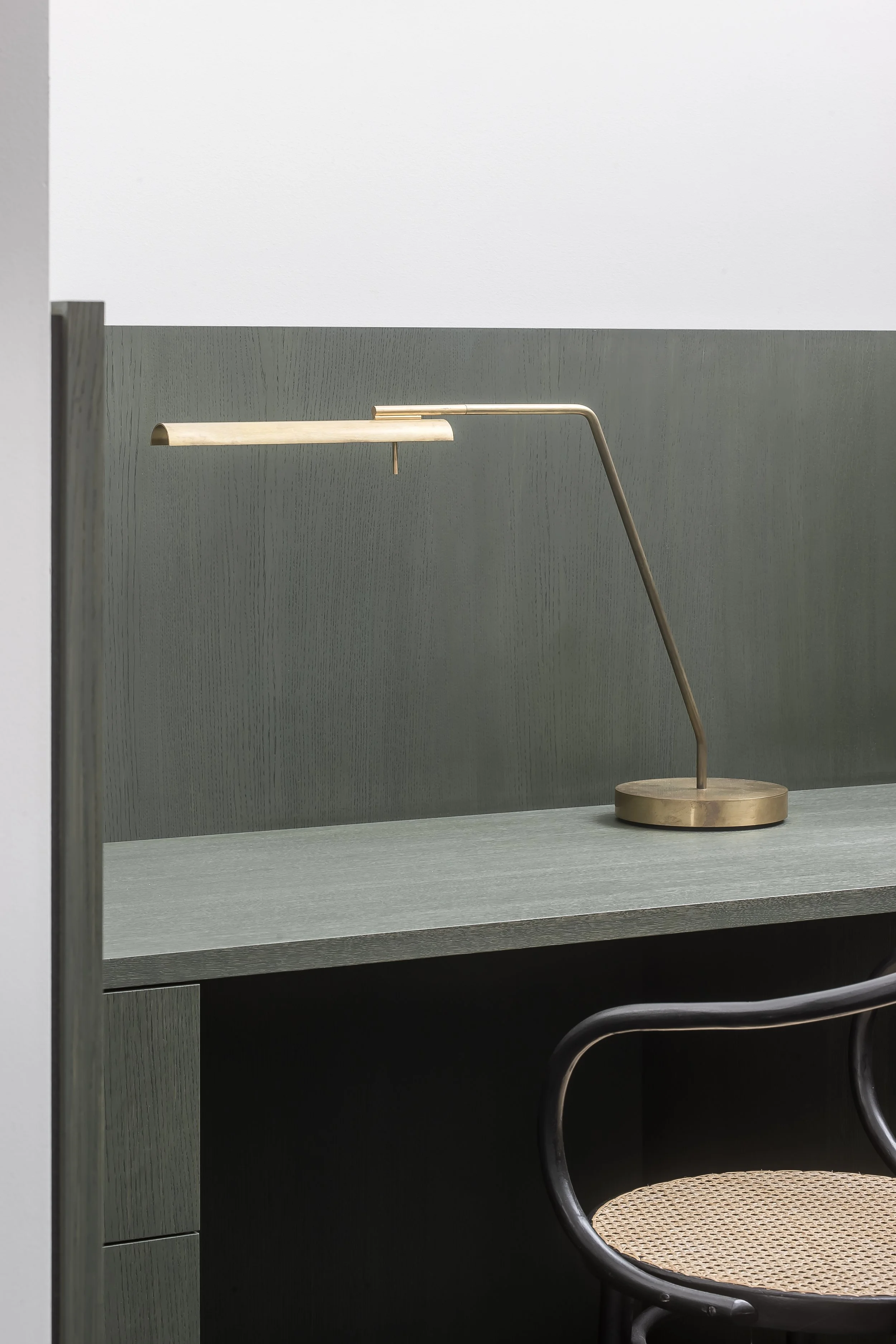 A minimalist desk setup with a modern lamp and a round woven chair in front of a gray wall.