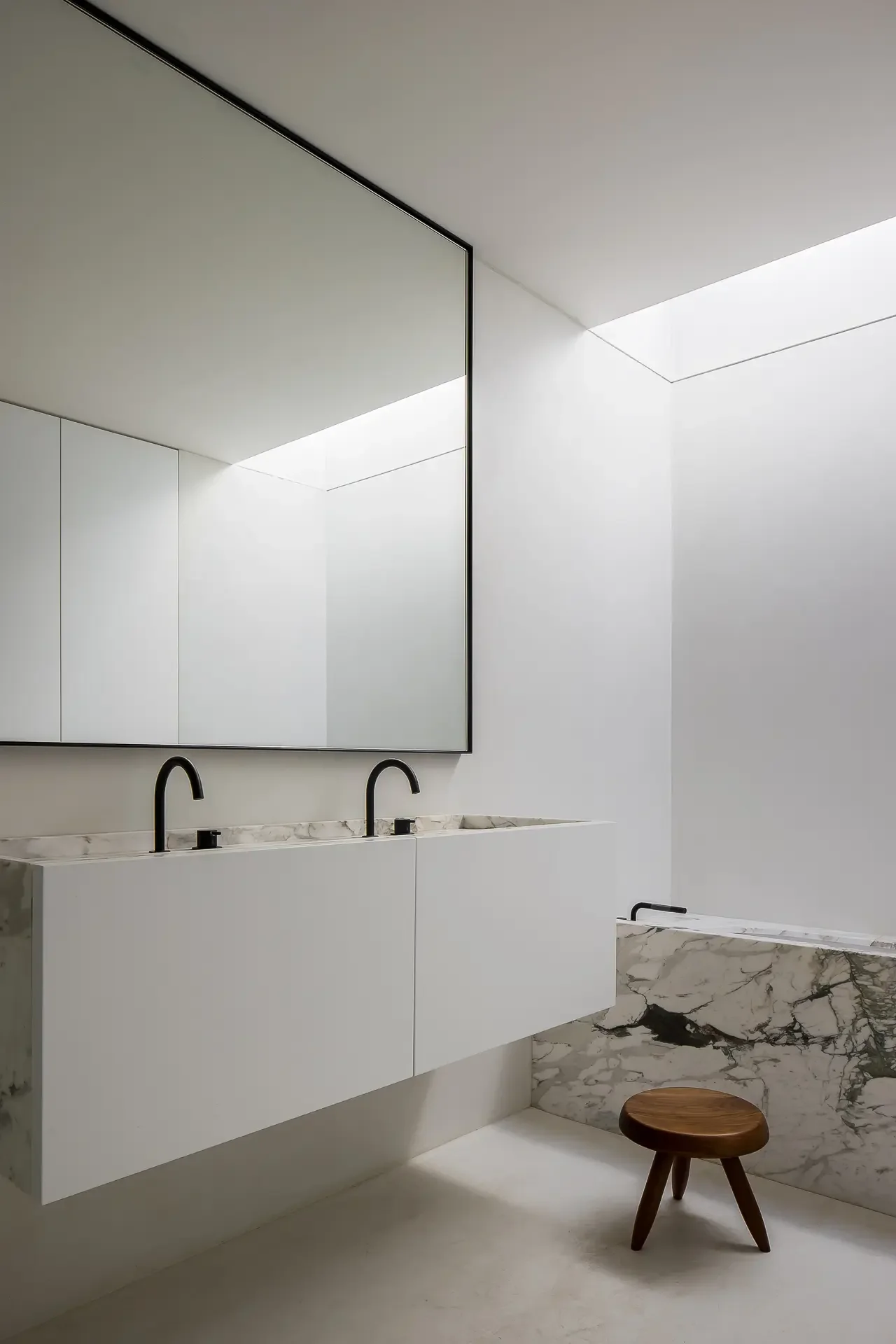 A minimalist bathroom with a large mirror, two black sinks with gold fixtures, white cabinetry, a marble countertop, a small wooden stool, and a marble accent wall. There is a skylight providing natural light.
