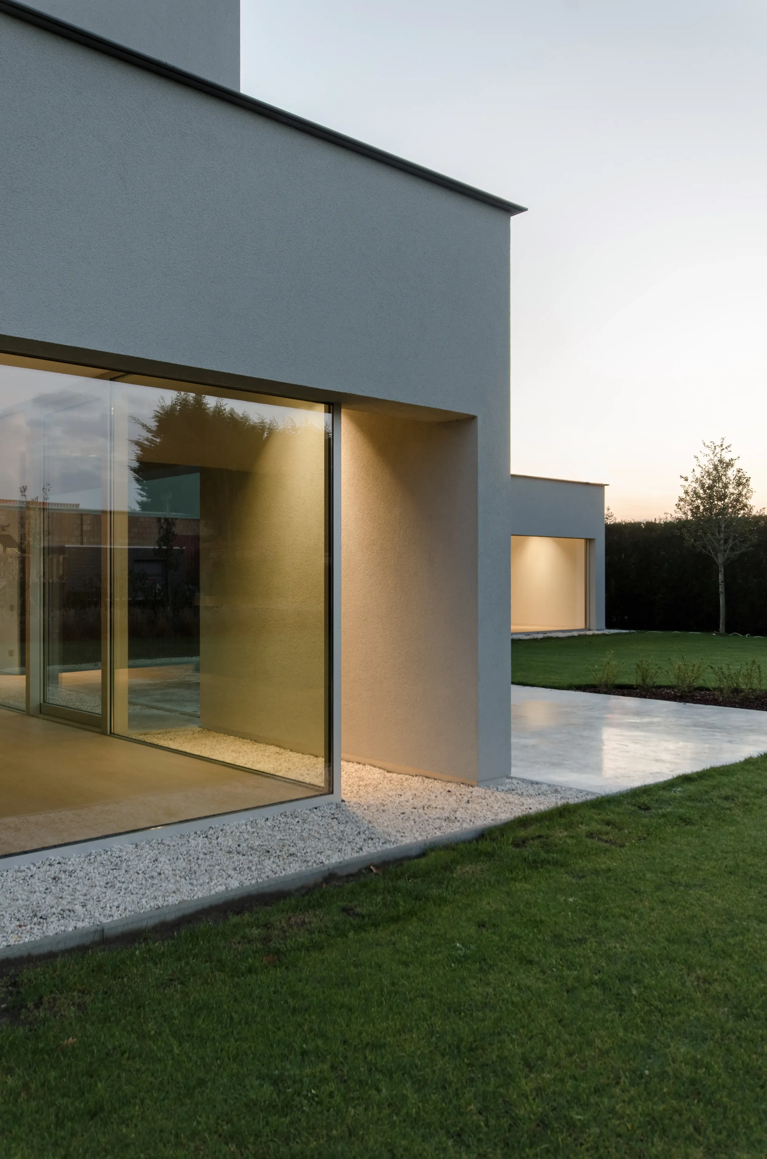 Modern white house with large glass windows and a lush green lawn at sunset.