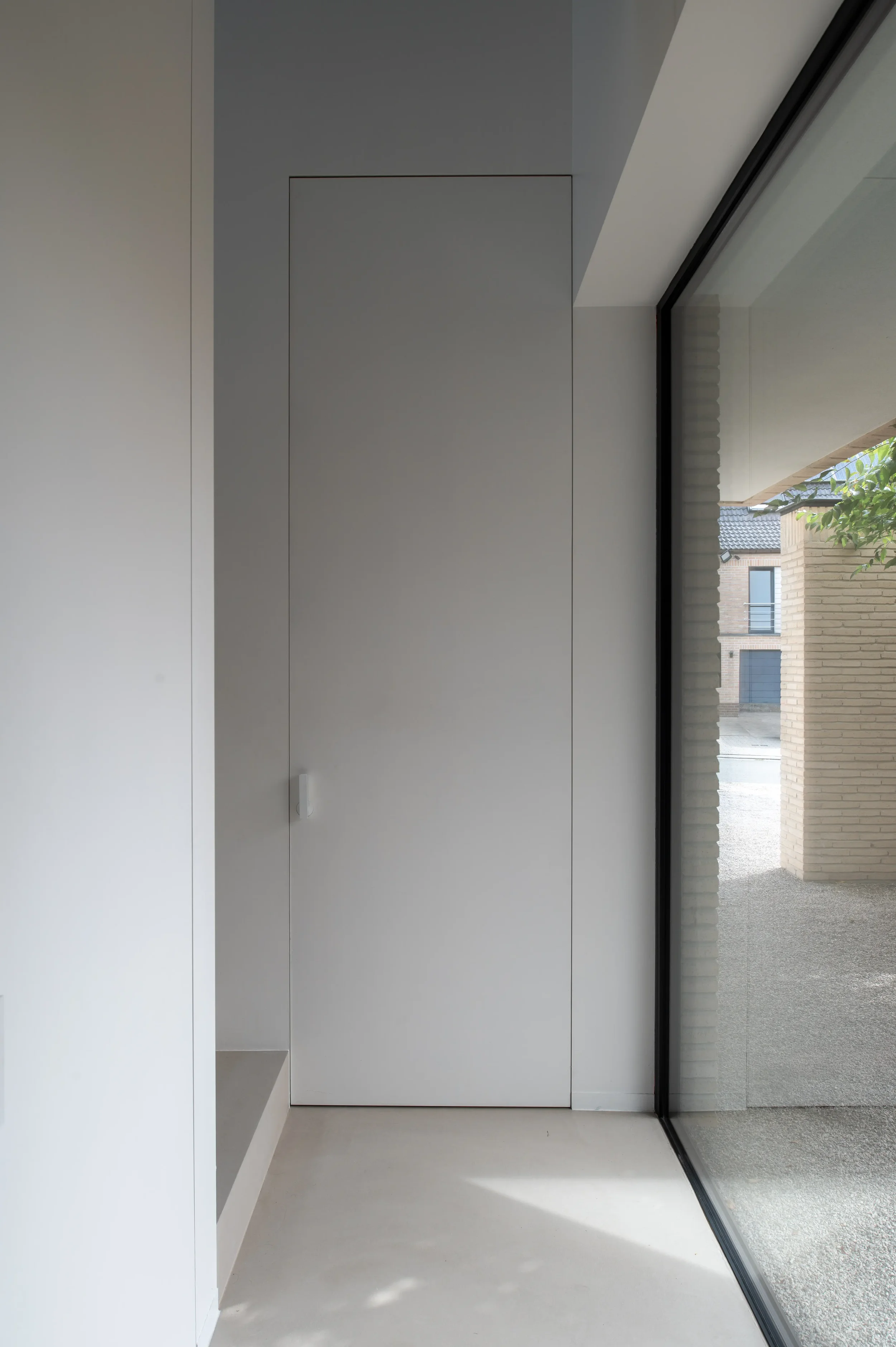 Interior space with white walls, a closed white door, and large floor-to-ceiling window showing an outside view of a building, trees, and blue sky.
