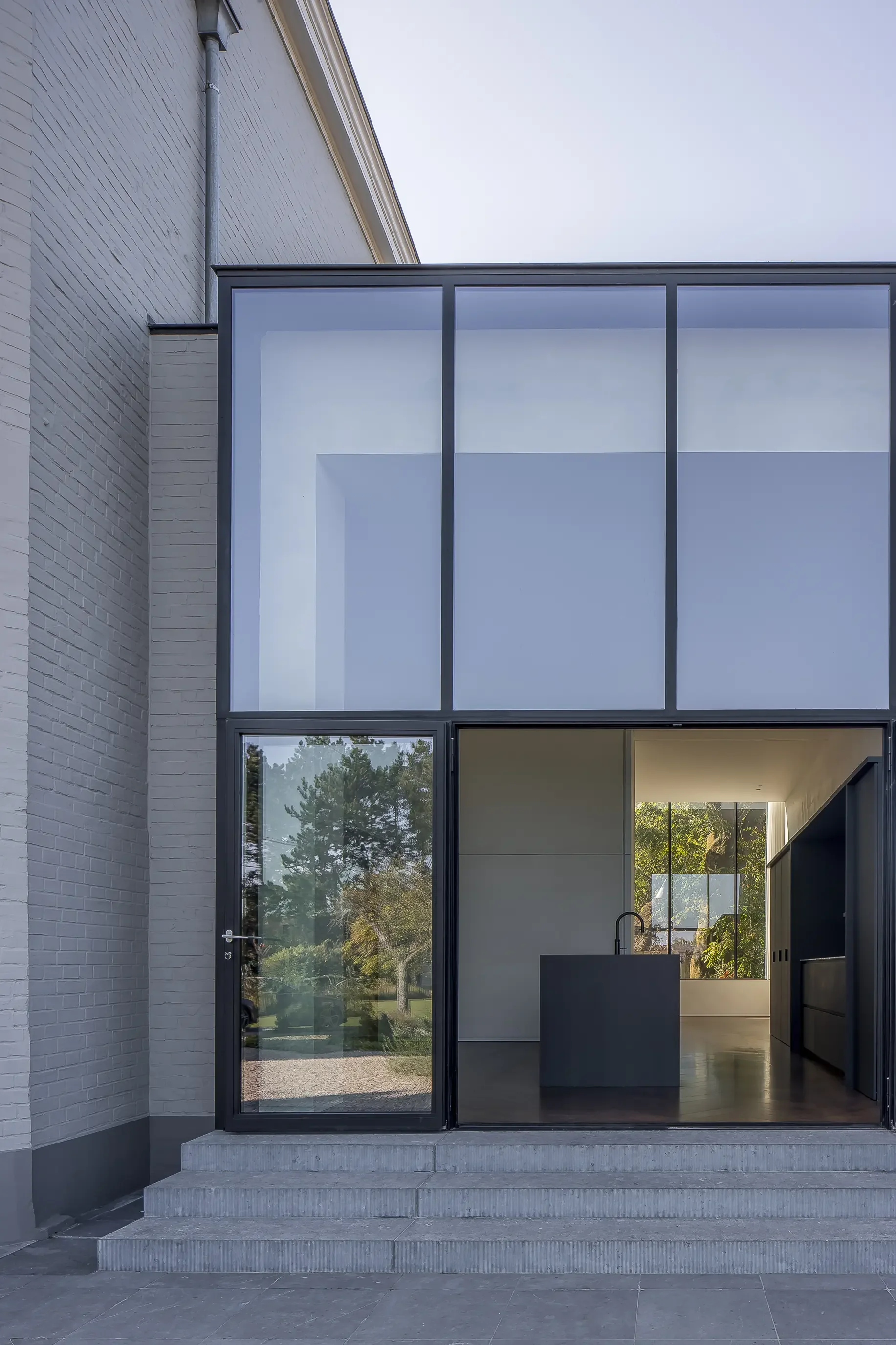 Modern house garden entrance with gray stairs, glass door, and large windows showing an interior kitchen with a black faucet and thin black cabinets, surrounded by white brick walls and trees outside.
