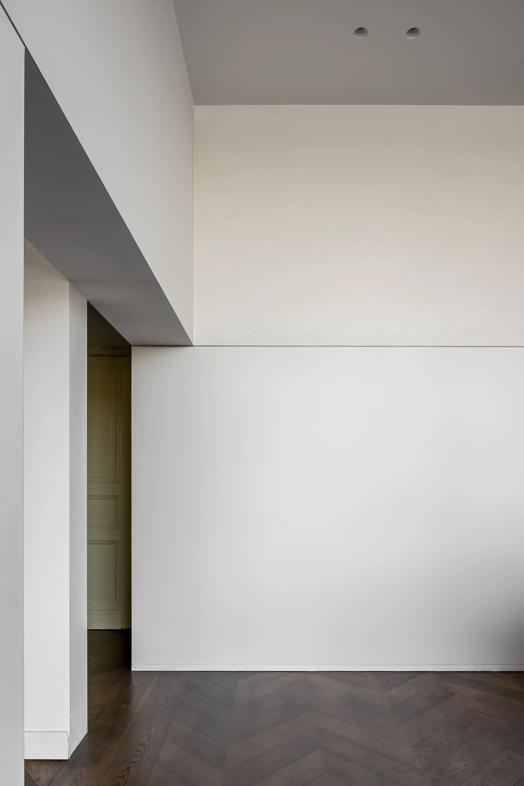 An empty white room with a high ceiling, featuring two small circular openings, a large white wall, a doorway partially visible on the left, and dark wooden herringbone-patterned flooring.