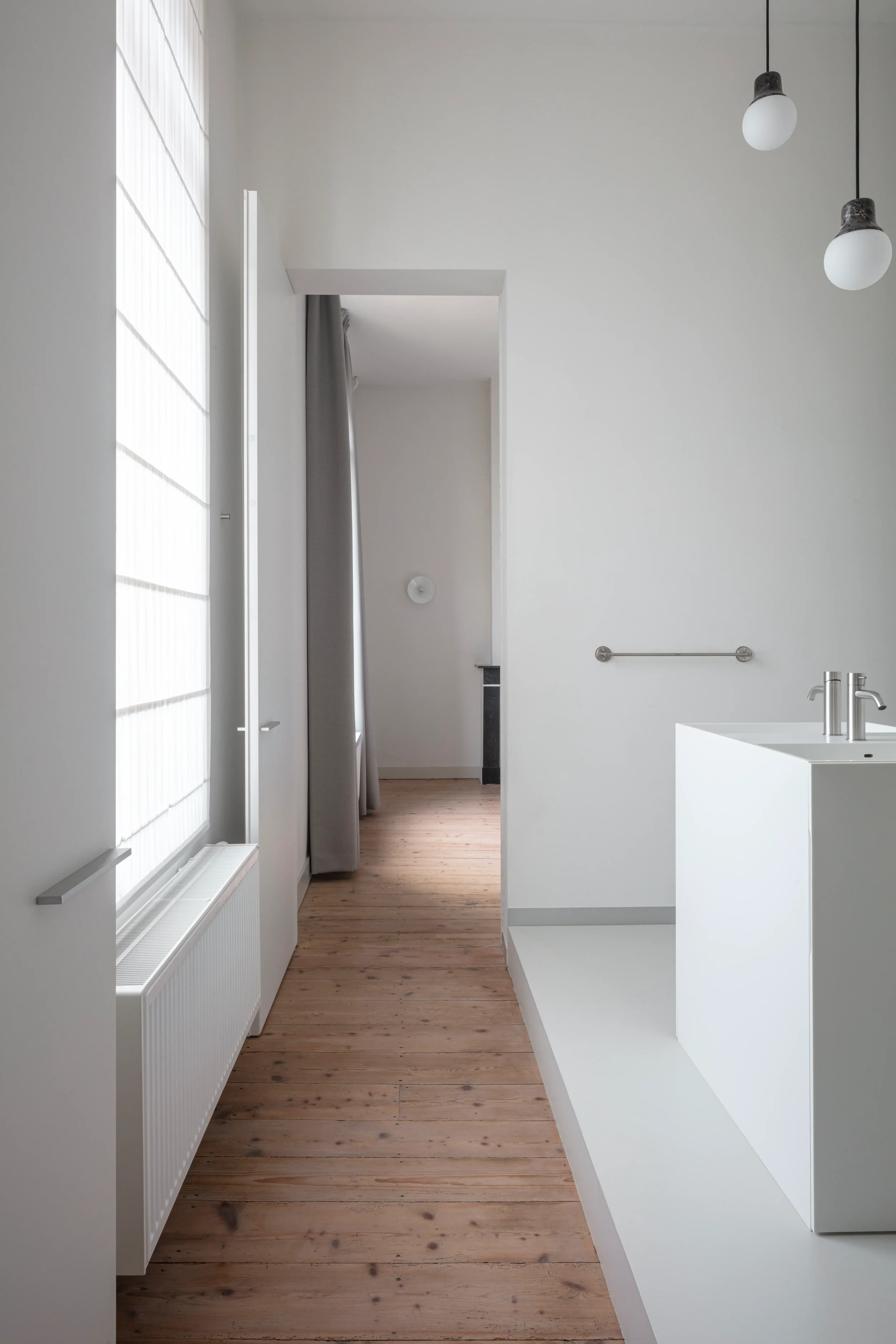 A minimalistic interior space with white walls, a wooden floor, a white sink with a silver faucet, modern light fixtures hanging from the ceiling, a silver towel bar, a radiator, and a doorway with a curtain.