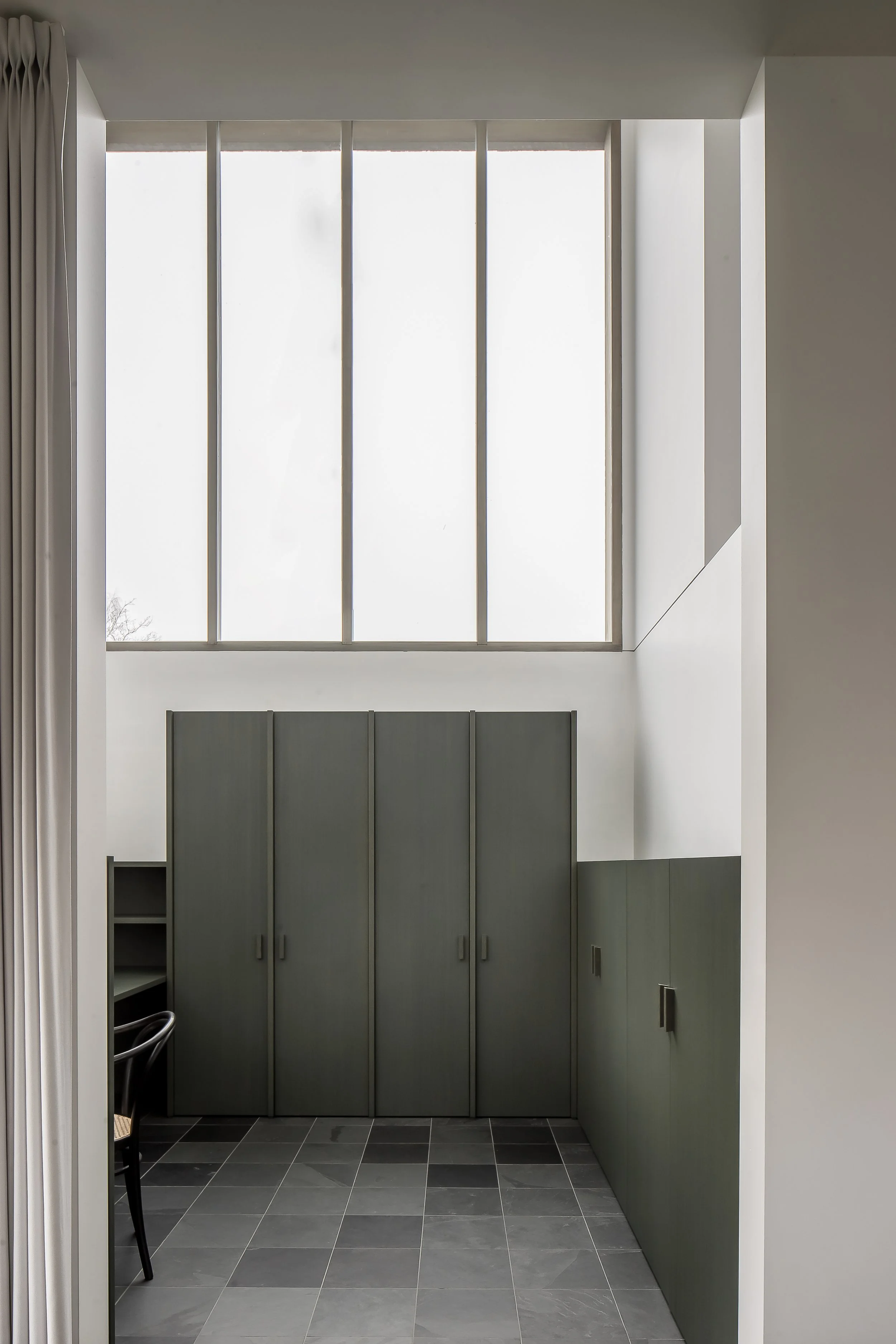 A large window with high glass panels and metal framing, allowing natural light into the room. To the left, a curtain is partially visible, and below, there are gray cabinets and a small, open shelving unit. The floor is covered with dark gray tiles.