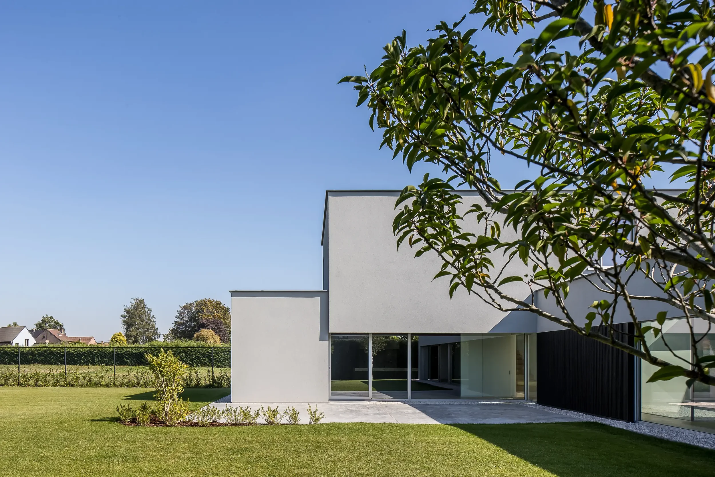 Modern white house with large sliding glass doors and a flat roof, set in a green yard with a clear blue sky overhead.