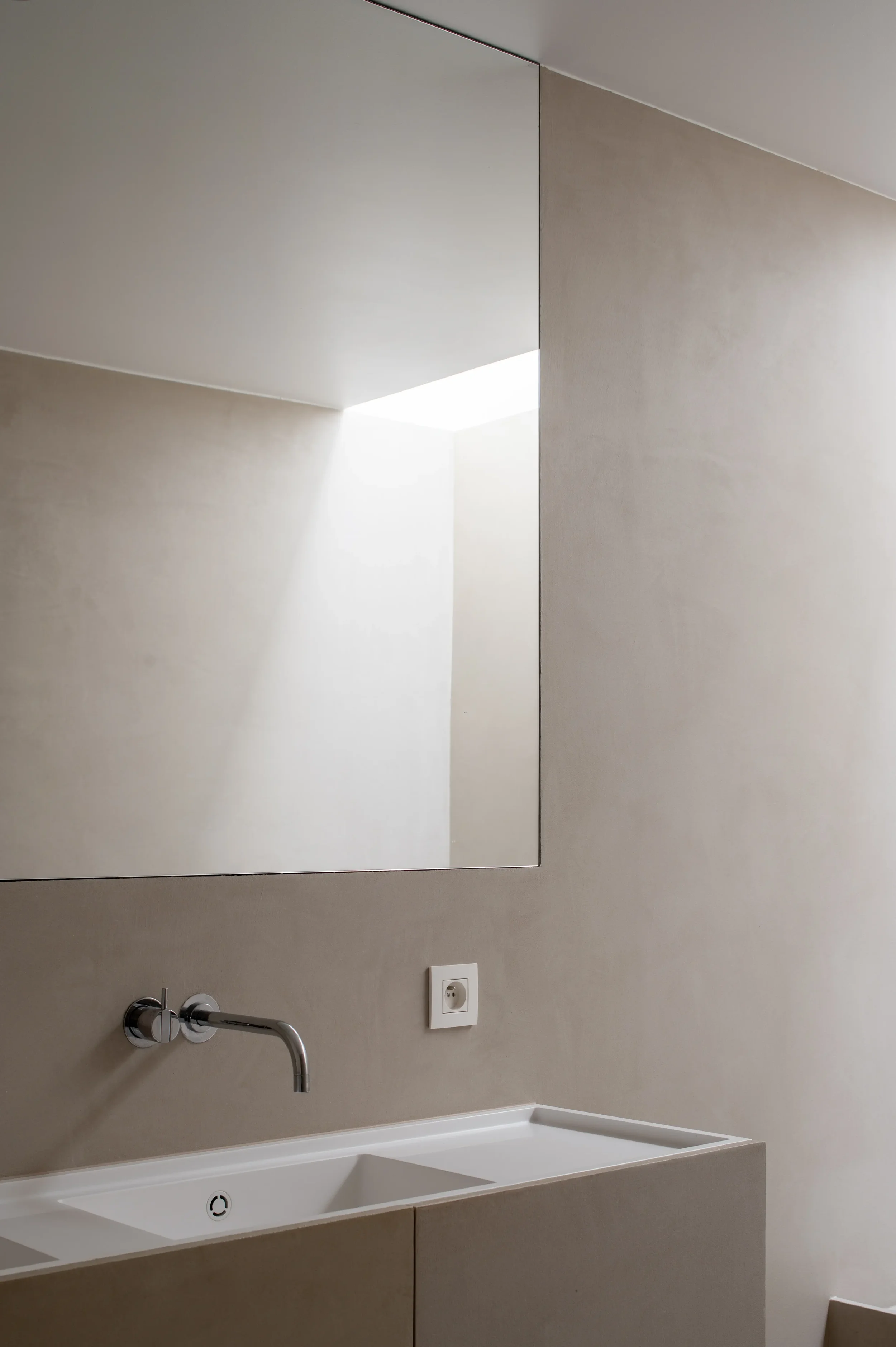 A bathroom with a beige wall, a large rectangular mirror, a white sink with a silver faucet, a double electrical outlet, and a wall-mounted soap dispenser.