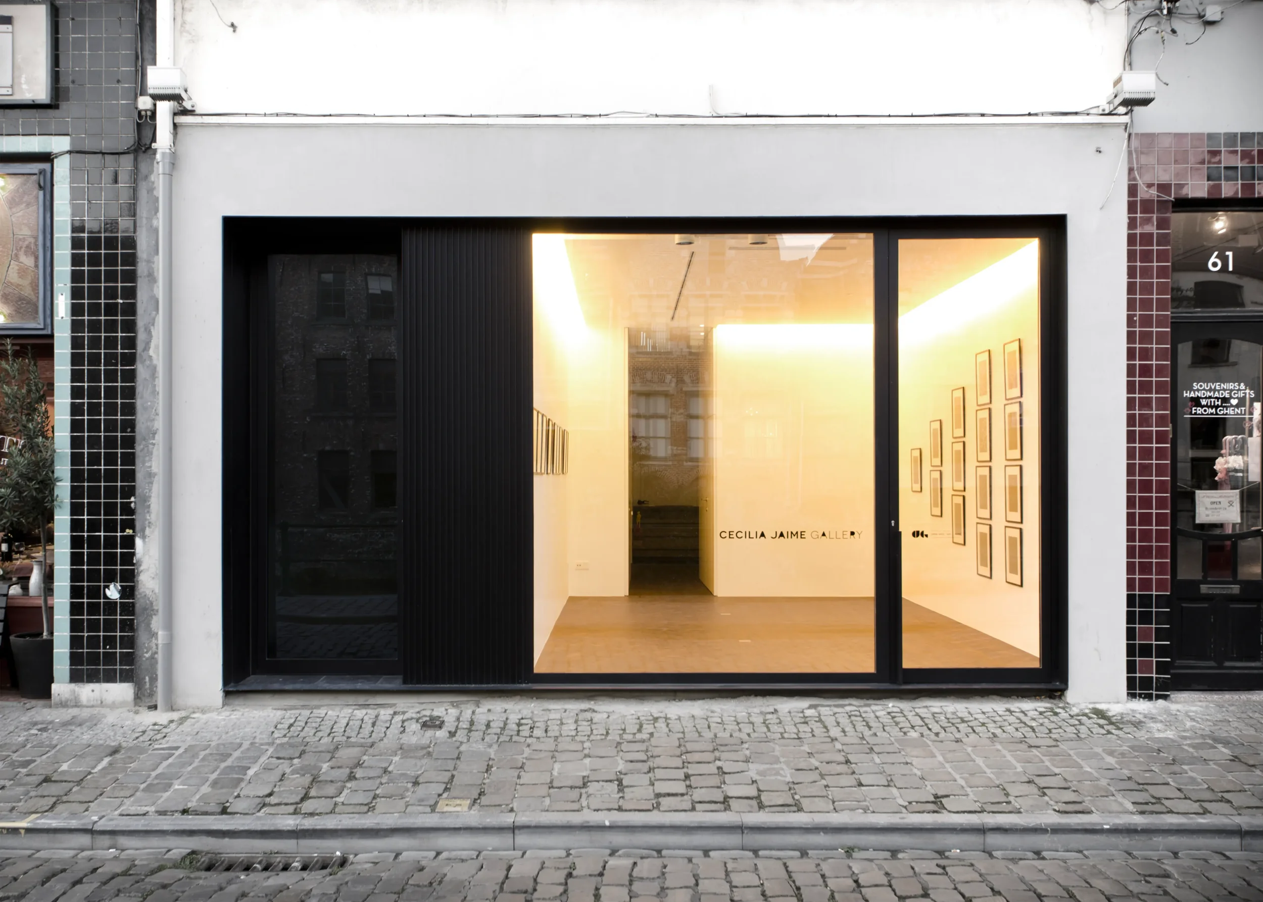 The storefront of Cecilia Jaime Gallery features a black entrance door and window, with the gallery's name displayed on the glass. Inside, framed artworks are visible on the wall, and a large illuminated ceiling light provides warm lighting.