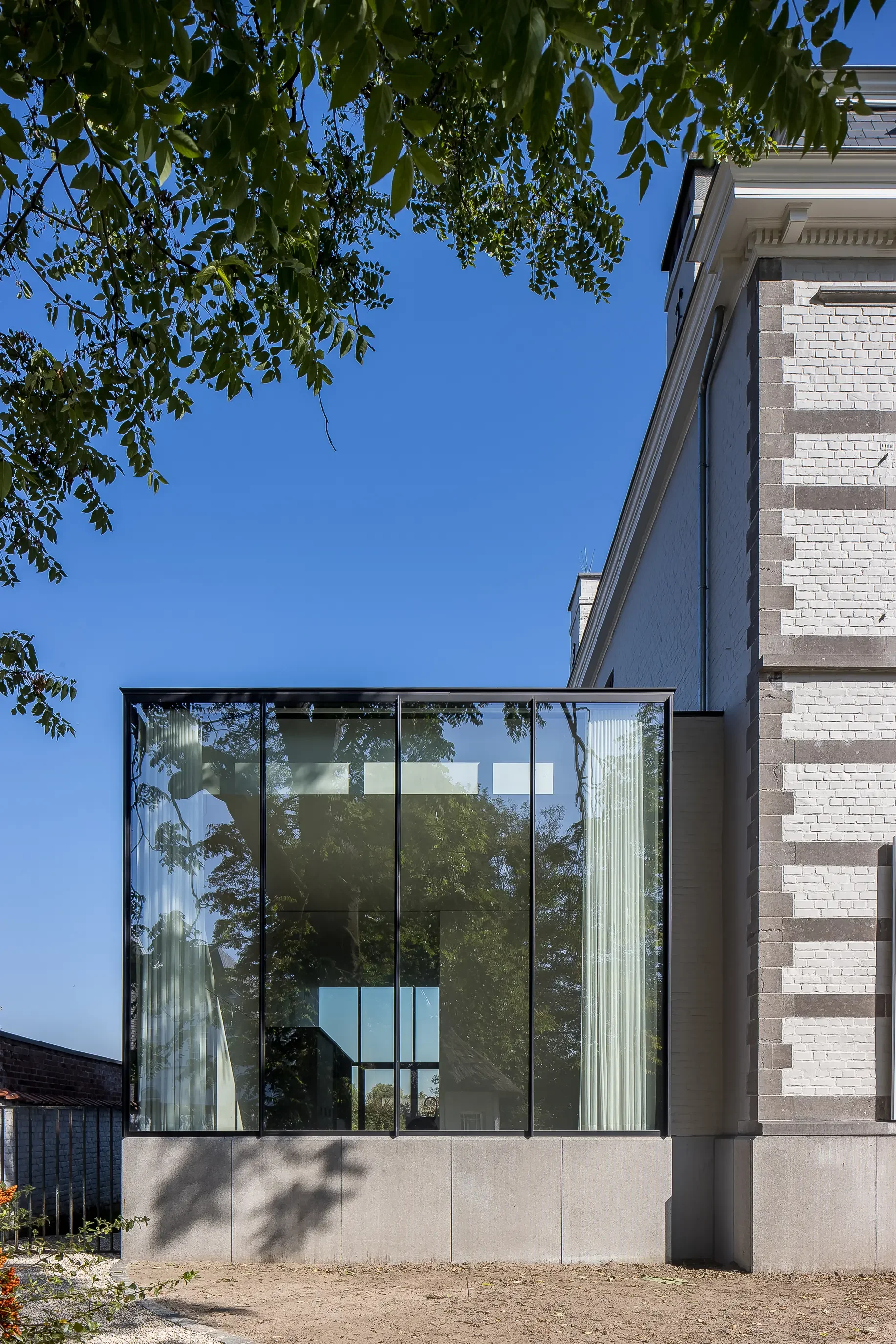 A modern extension of a classic house with an exterior made of white bricks, large glass windows with curtains, a rooftop, and a tree with green leaves in the foreground.