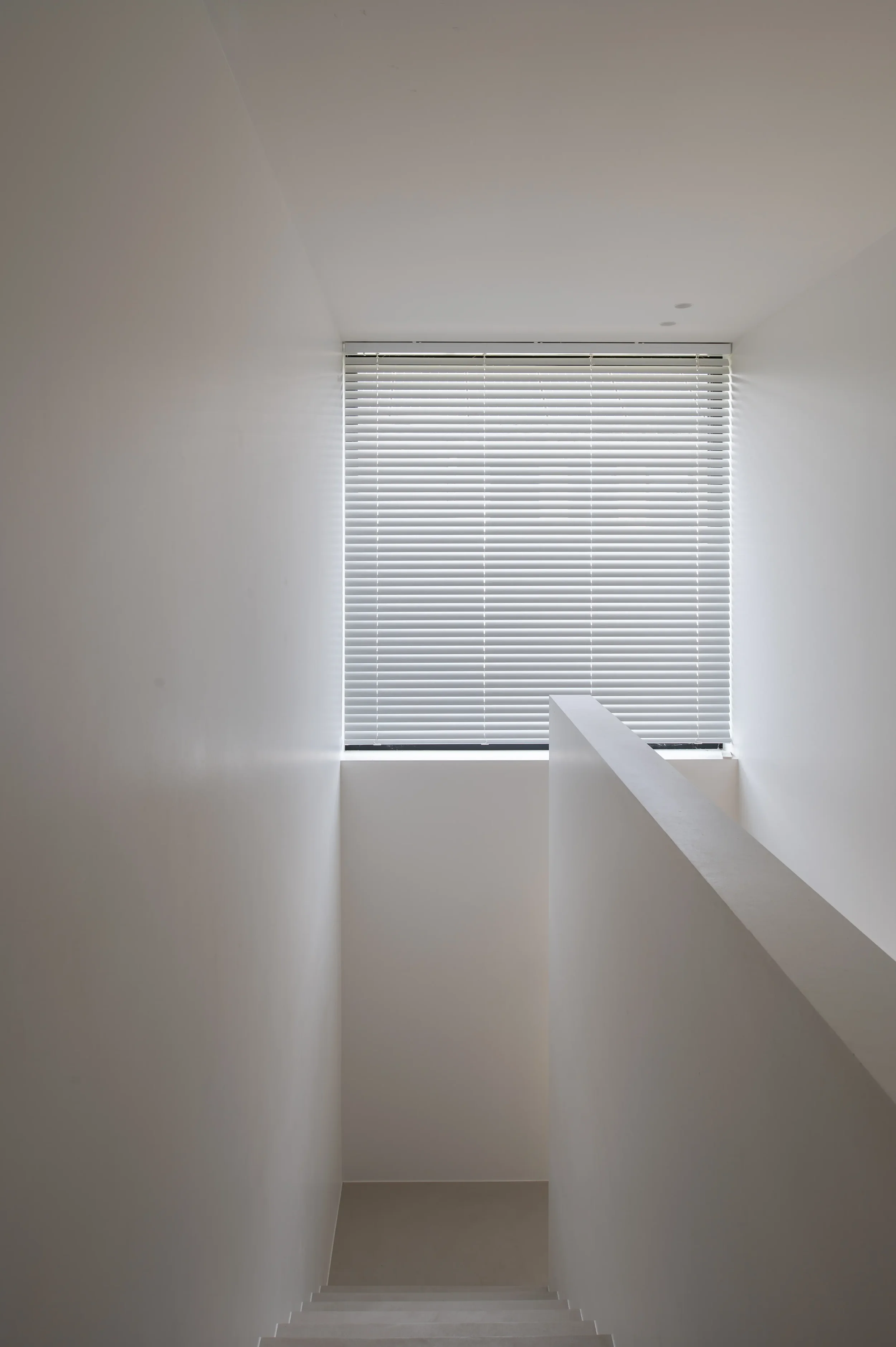 Bright corridor with white walls, modern staircase, and a large window with white blinds at the end.