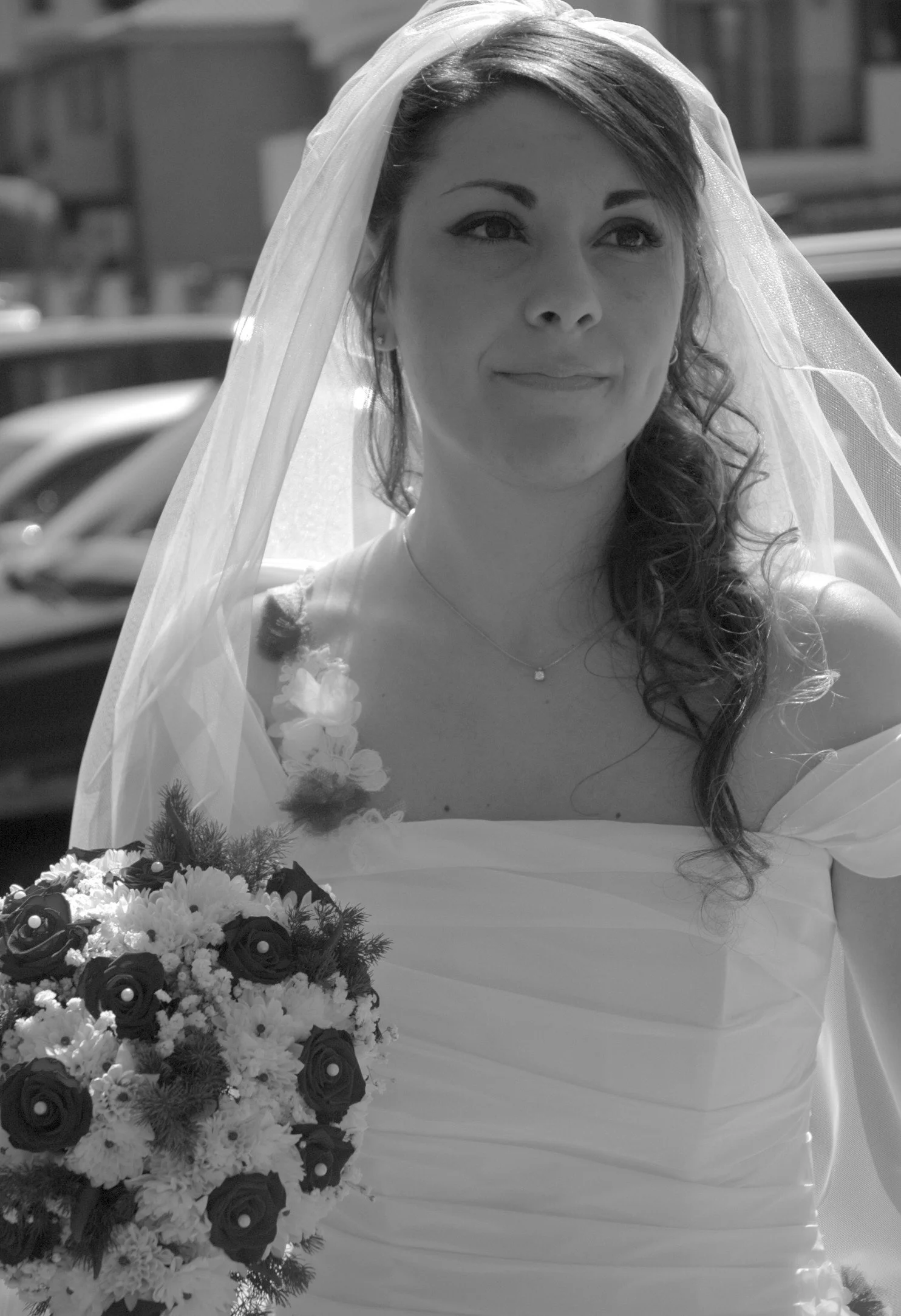 Una donna sorridente con abito bianco da sposa e veletta posando con un bouquet di fiori davanti a uno sfondo di veicoli parcheggiati.