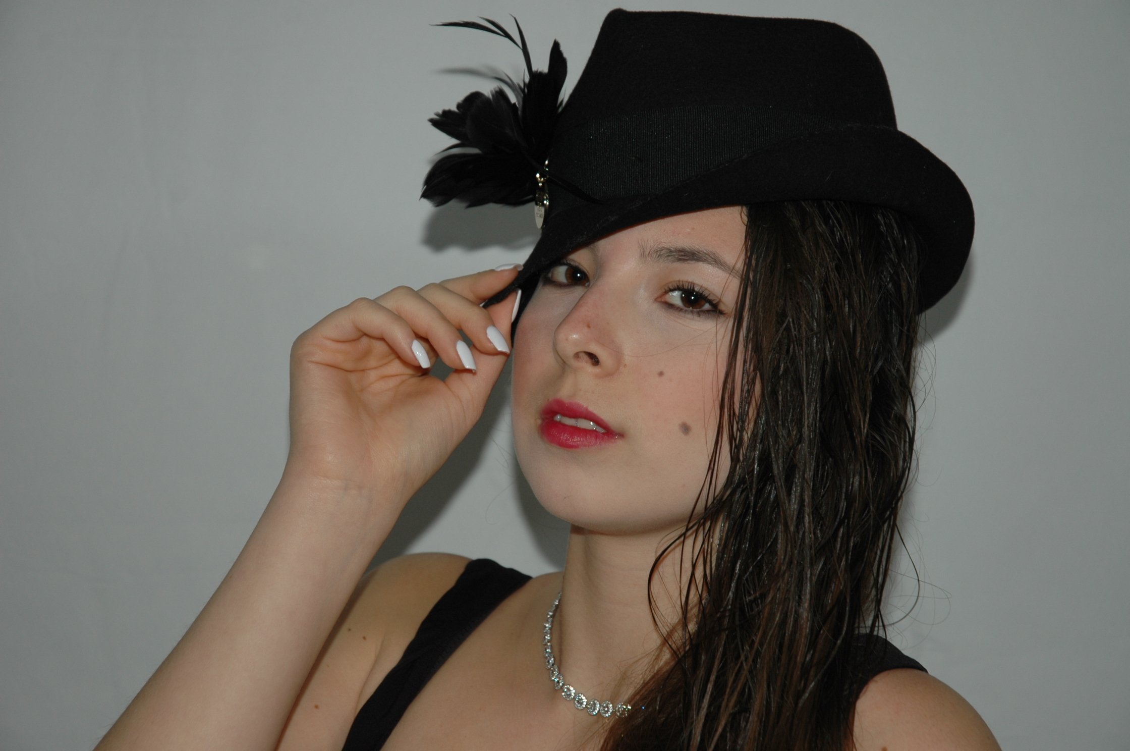 Ragazza con capelli ricci scuri indossa una bombetta nera con piume e adesivo, occhi chiari, rossetto rosso, con collana di brillantini, sfondo grigio.