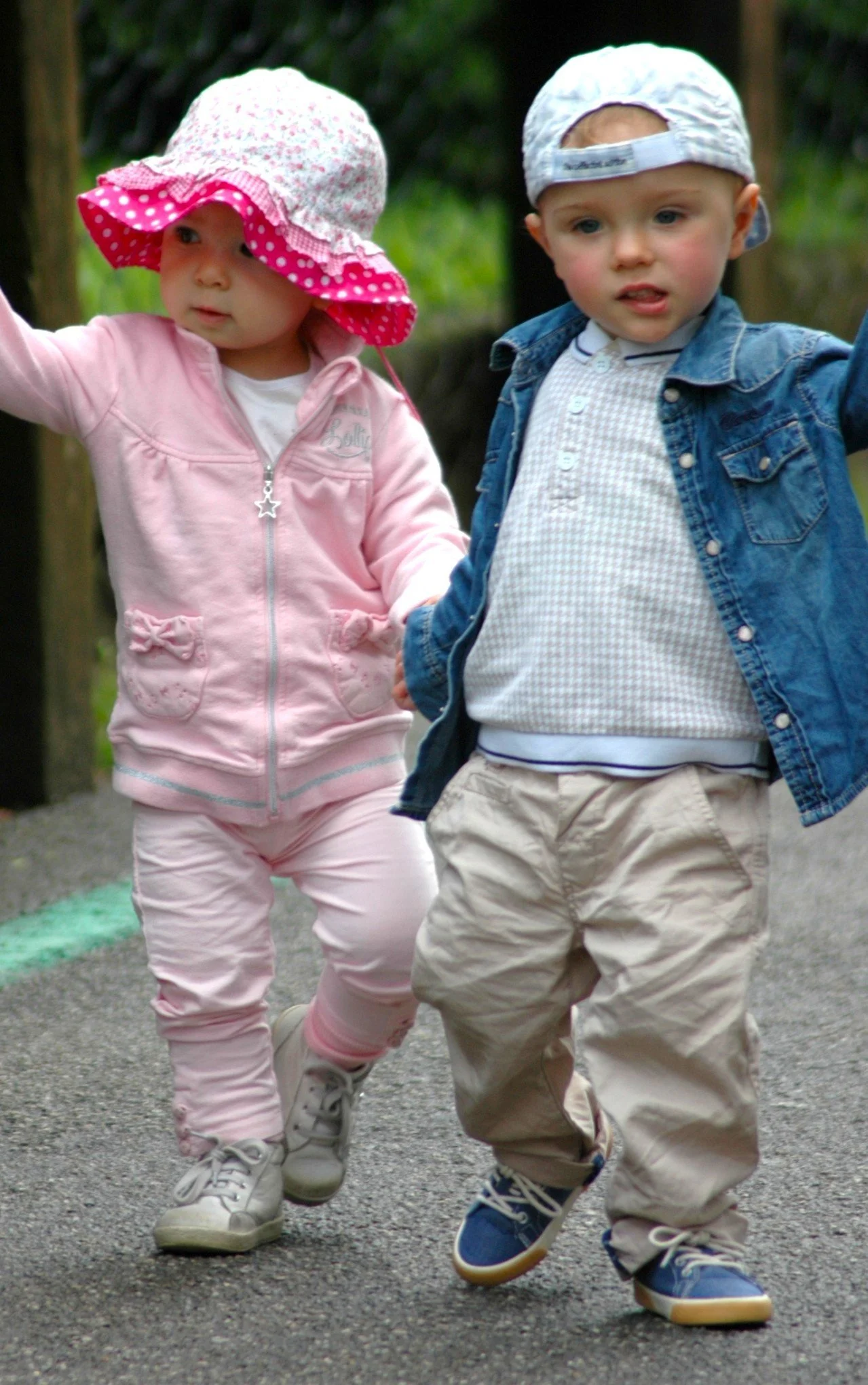 Due bambini piccoli, un ragazzo e una ragazza, mentre camminano insieme all'aperto, entrambi indossano cappelli e abbigliamento casual. La scena si svolge in un ambiente naturale, forse un parco.