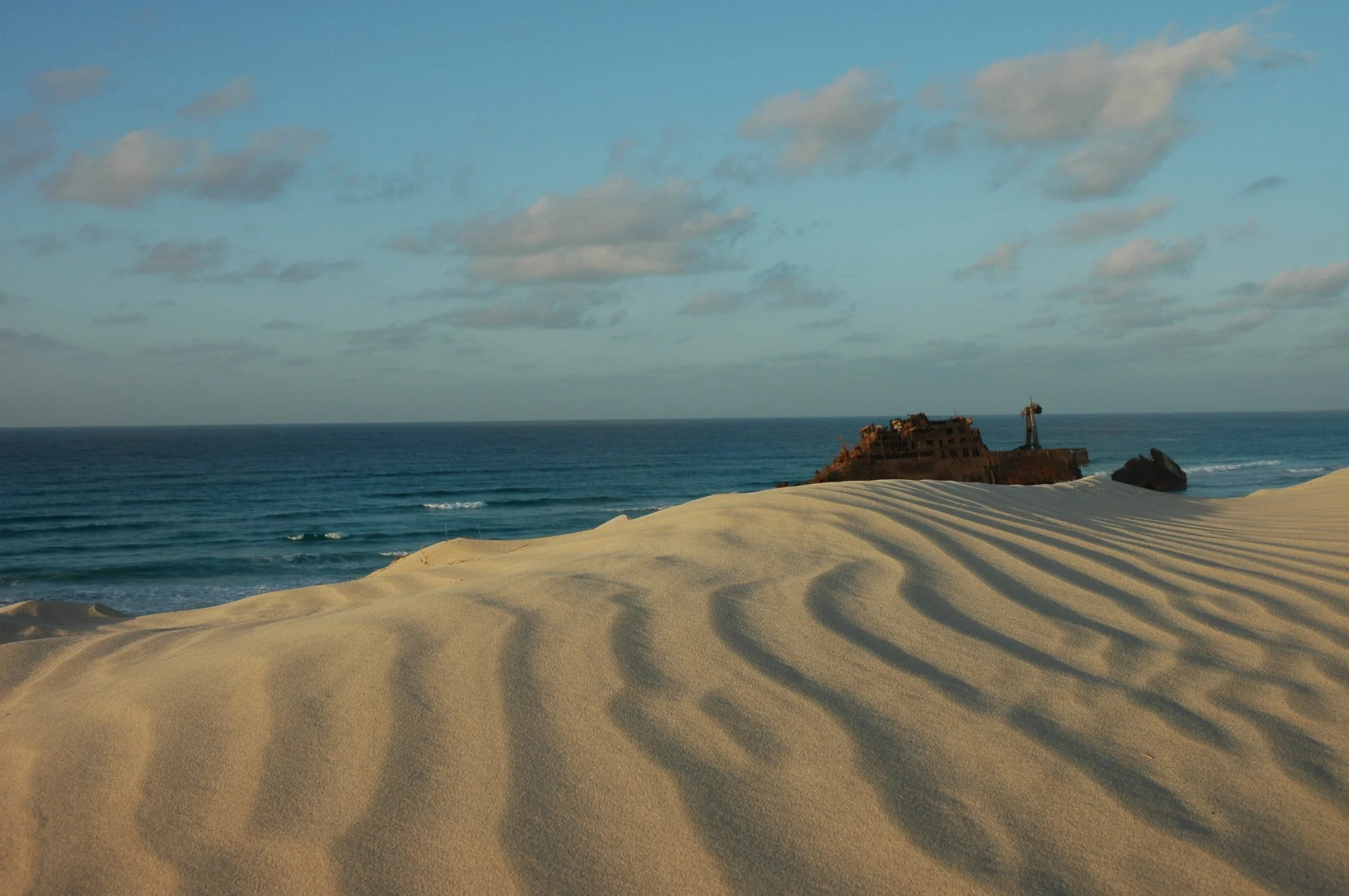 Spiaggia con dune di sabbia dorata, mare calmo e un relitto di nave vicino alla riva, con un faro sulla roccia a lato.