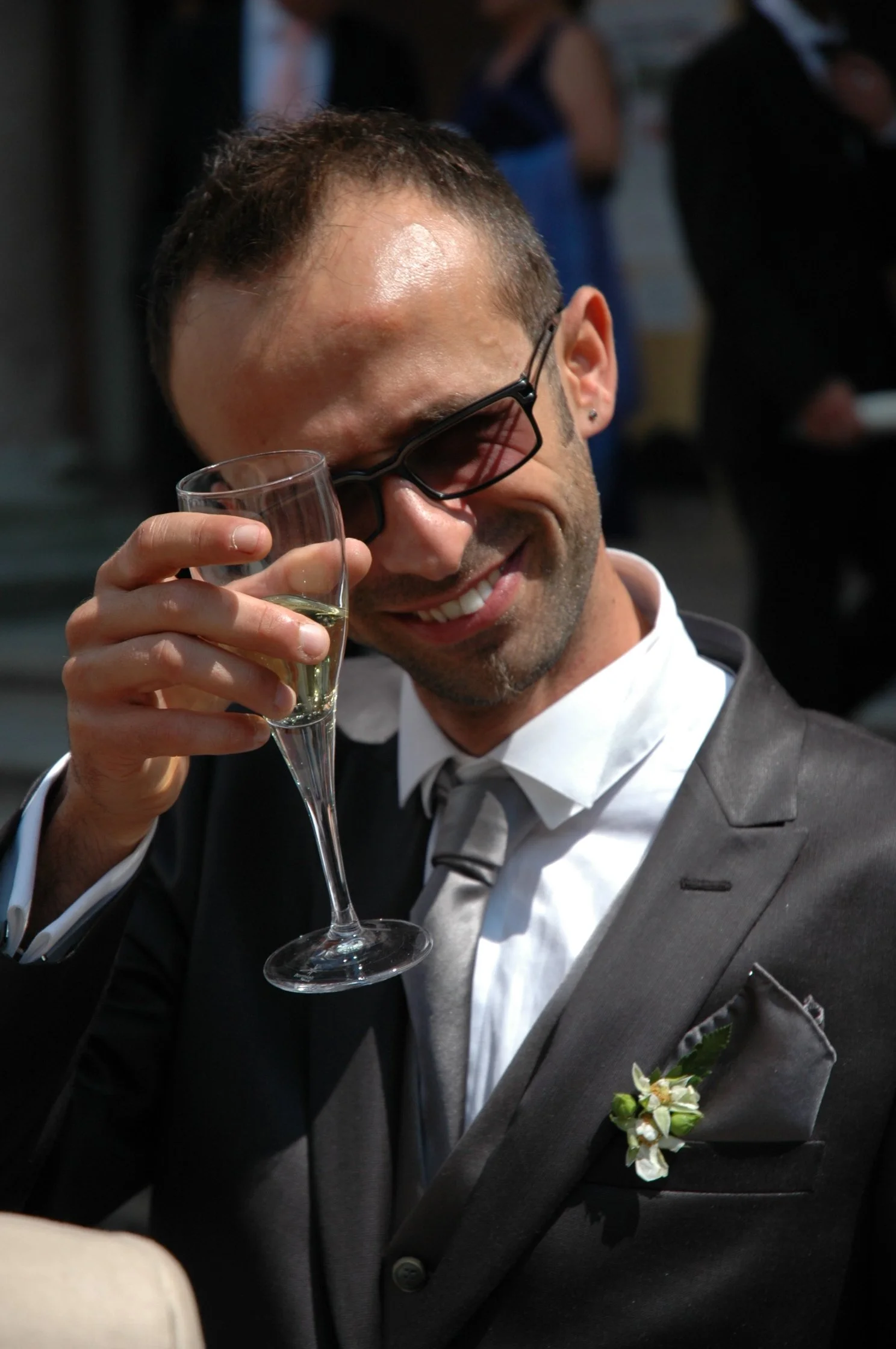 Uomo sorridente con occhiali scuri al matrimonio, che tiene un bicchiere di spumante, indossa un abito grigio con fiore bianco sulla giacca.