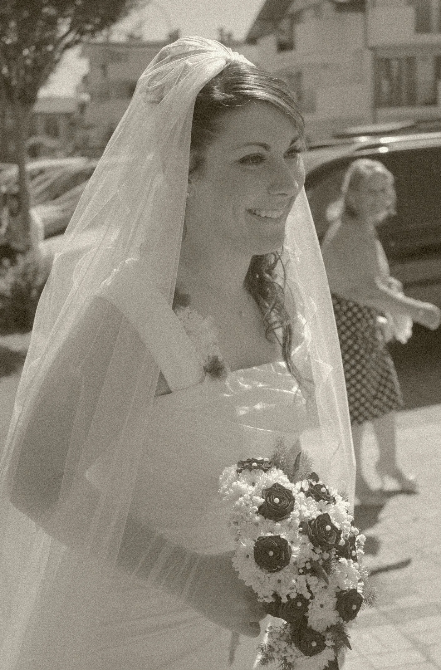 Una donna sorridente con un velo da sposa che tiene un mazzo di fiori, sullo sfondo ci sono altre persone e un edificio.