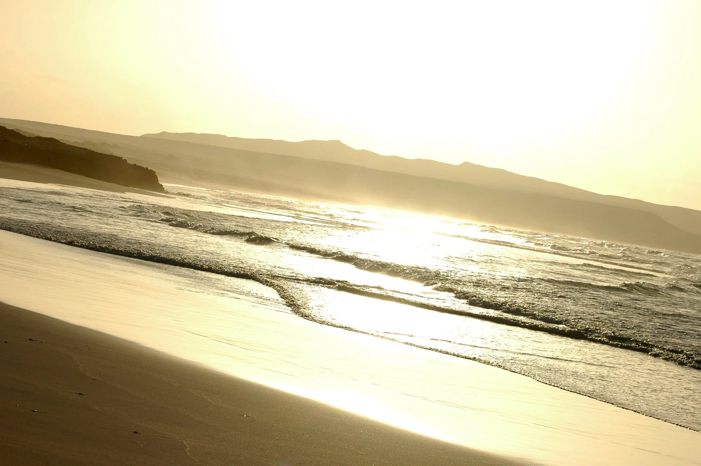 Spiaggia al tramonto con onde che si infrangono sulla riva e montagne sullo sfondo.