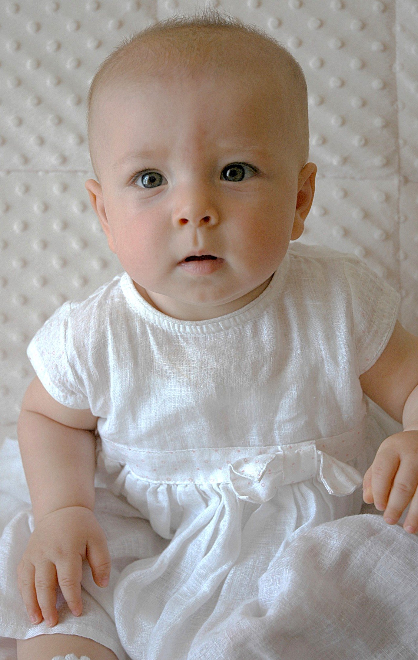 Ragazzo neonato con capelli corti e vestito bianco, seduto su uno sfondo chiaro con pattern a rilievo.