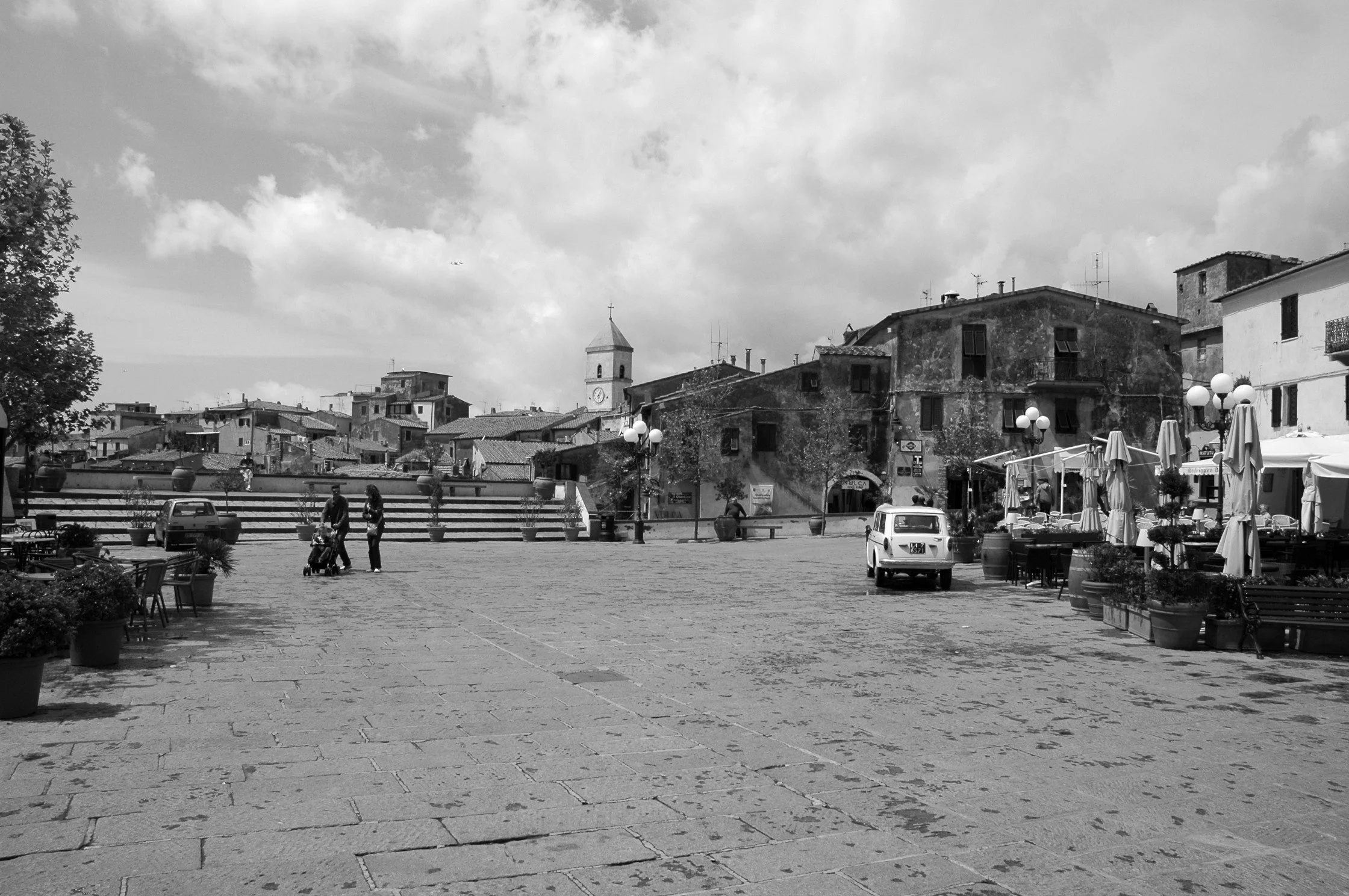 Piazza di un villaggio con edifici storici, campanile, persone che camminano, auto parcheggiate e tavoli all'aperto con ombrelloni, in un giorno luminoso con nuvole nel cielo.