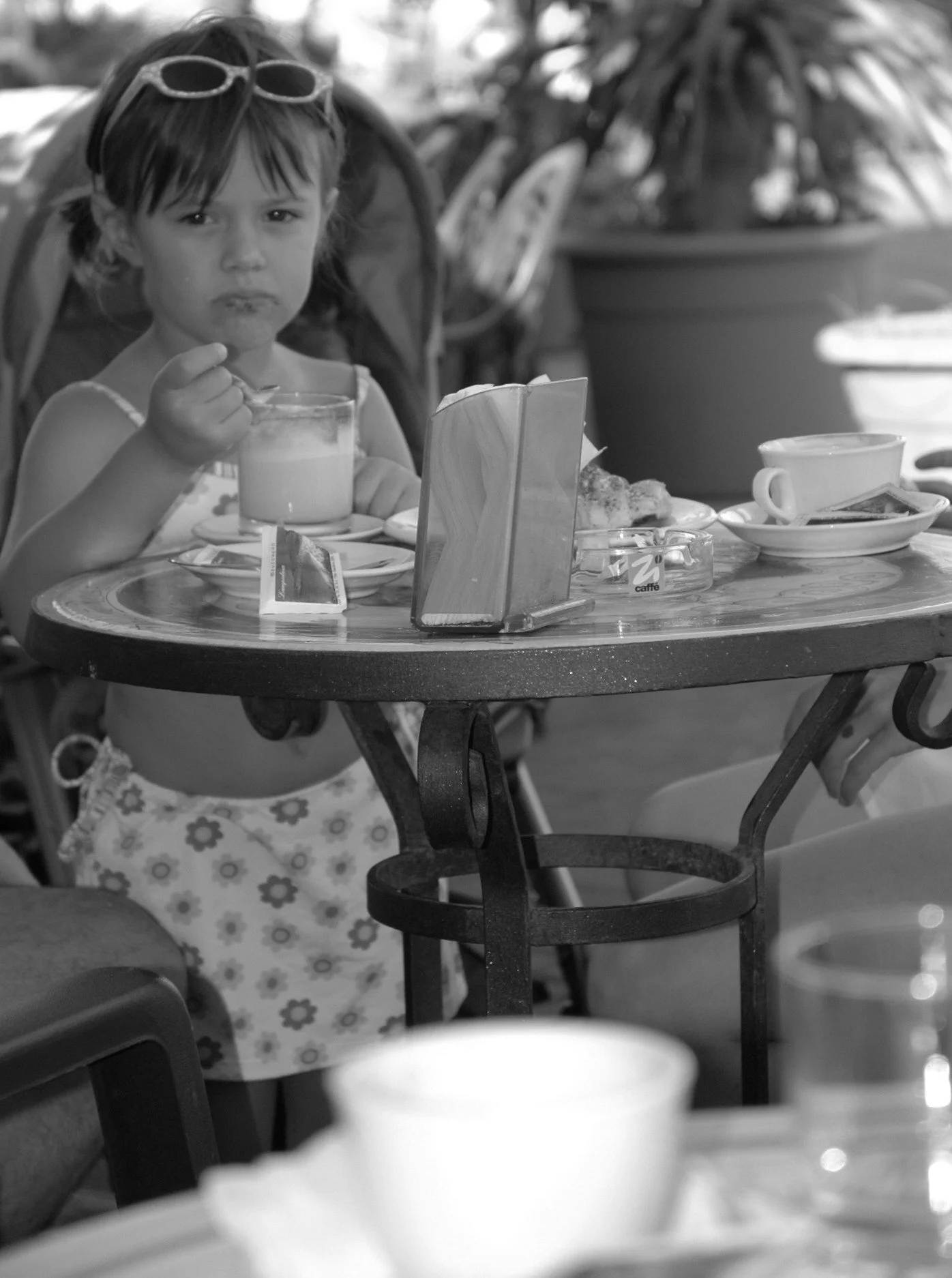 Una giovane ragazza con occhiali da sole sulla testa, seduta a un tavolo di un caffè o ristorante, che mangia una crema o gelato in un bicchiere. Sulla tavola ci sono anche alcune tazze e negozi di zucchero.