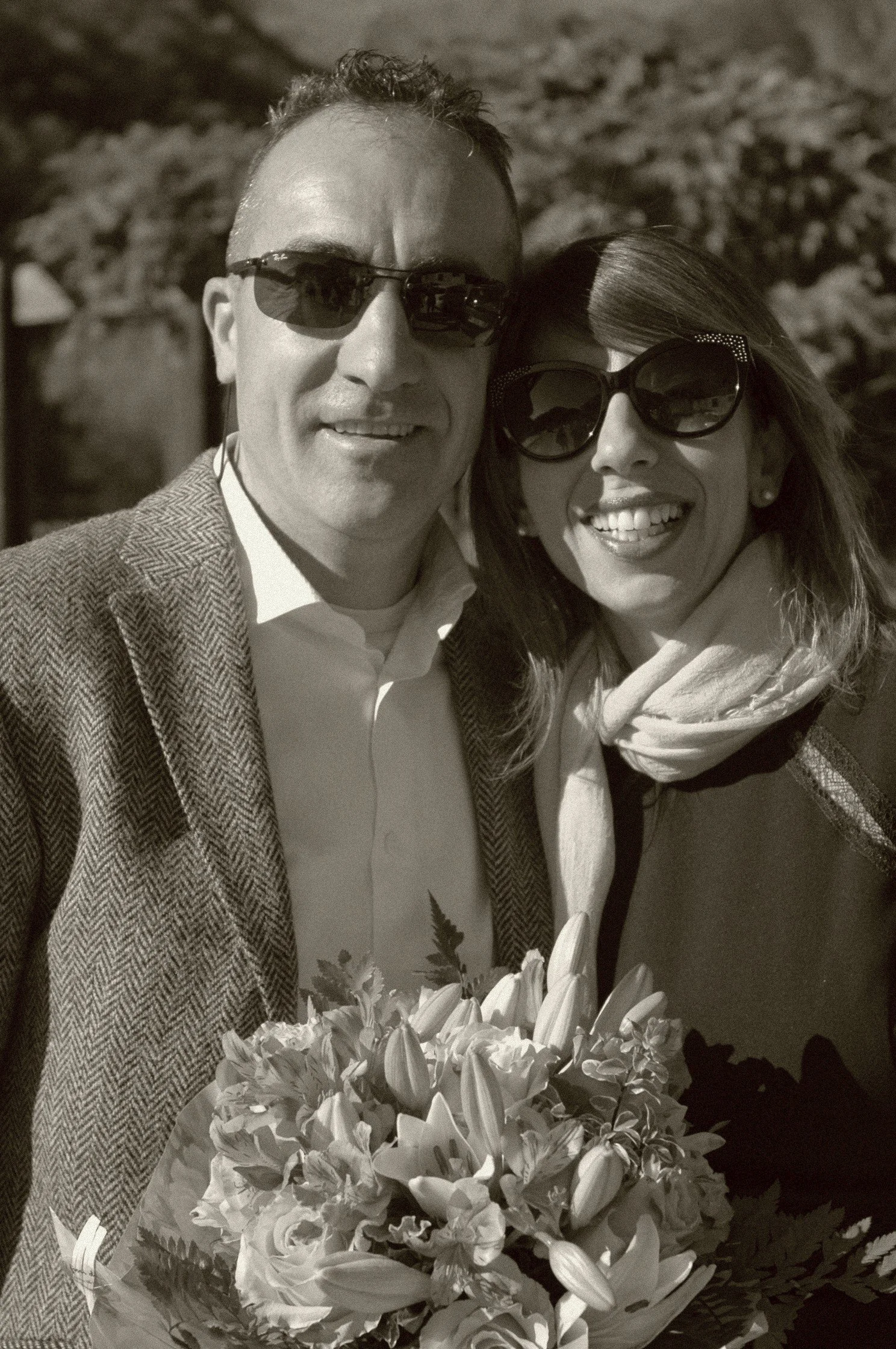 Una coppia sorridente con occhiali da sole si abbraccia e tiene un mazzo di fiori, in un ambiente dove ci sono alberi sullo sfondo.