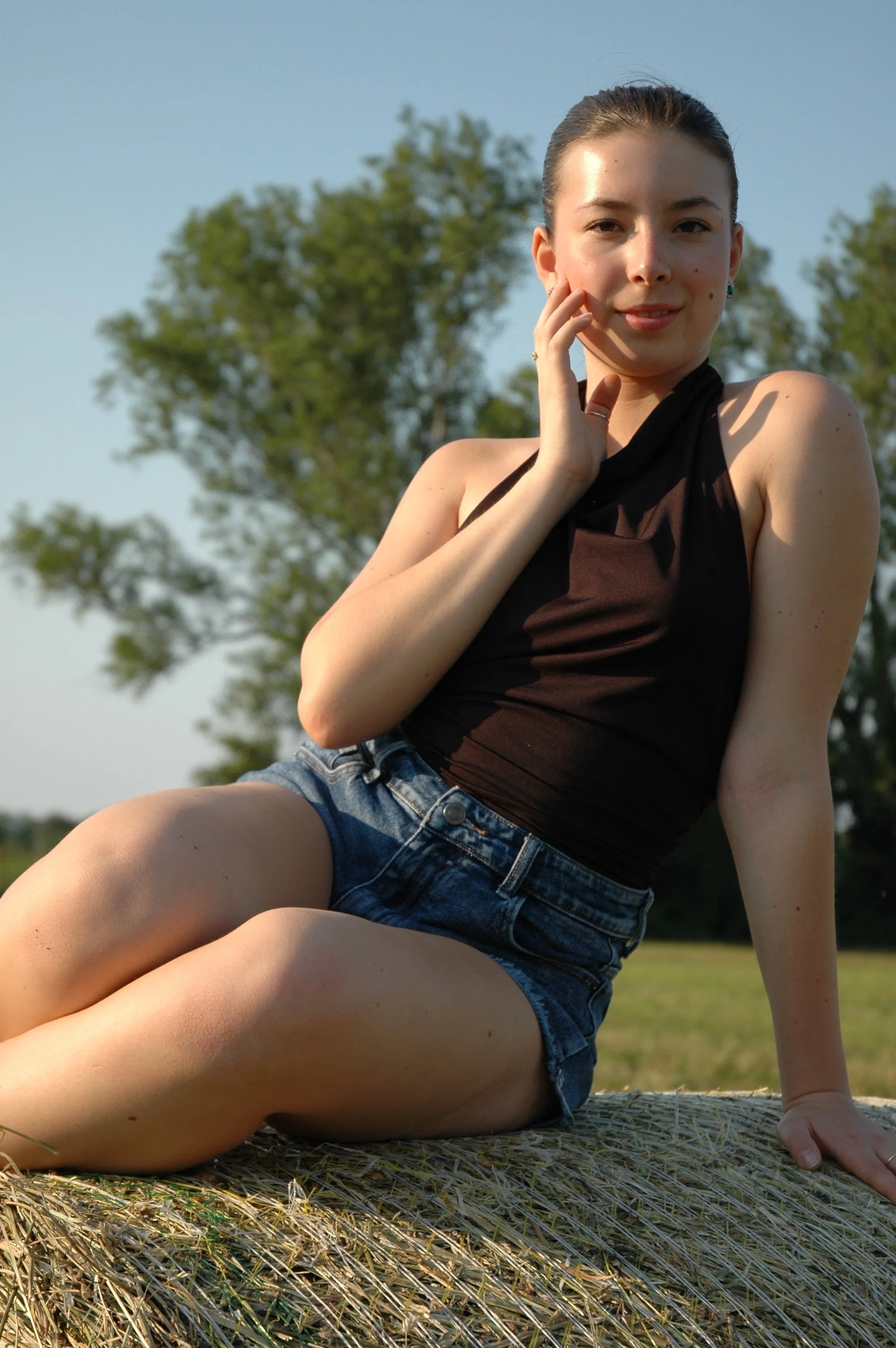 Ragazza seduta su un pagliaio in un campo verde, con albero sullo sfondo, indossa una maglietta nera e jeans corti, guarda sorridendo verso la fotocamera